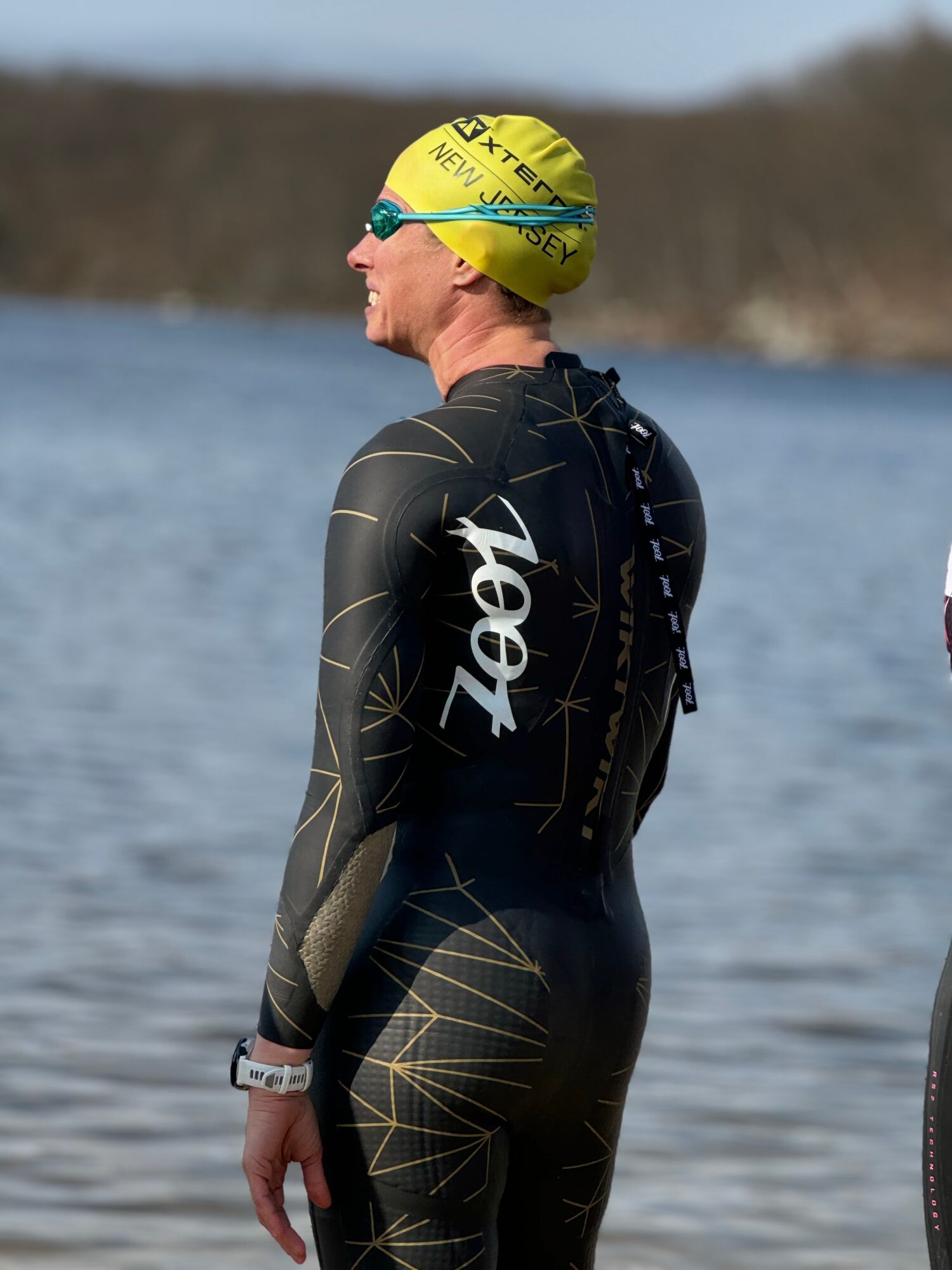 Person in a wetsuit and yellow swim cap standing by water, facing sideways, with a natural landscape background.