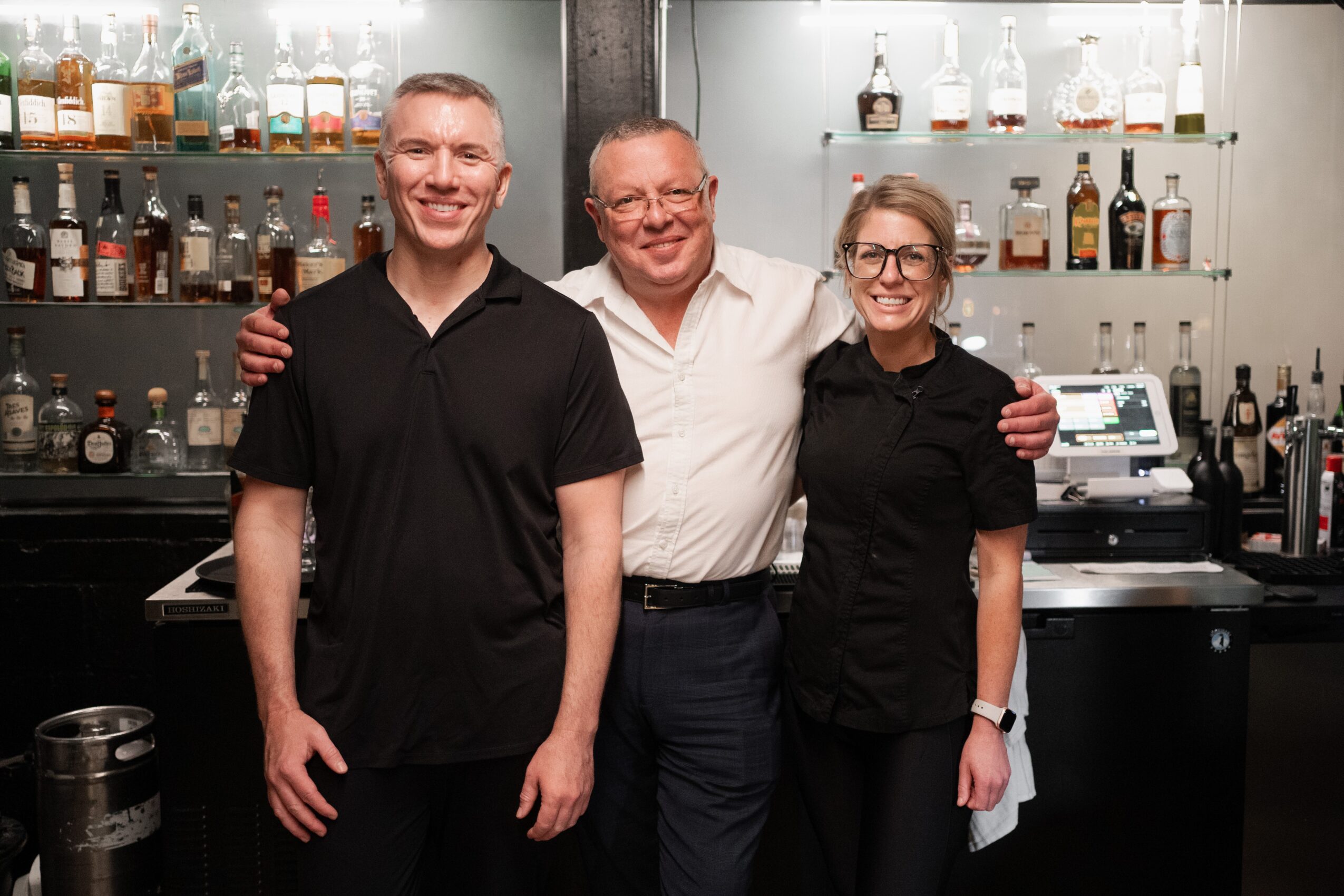 Three people standing behind a bar, smiling, with bottles and bar equipment in the background.
