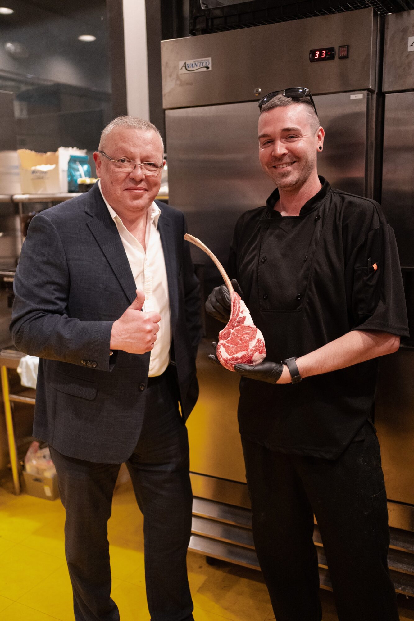 Two men standing in a kitchen, one in a suit giving a thumbs-up, the other in a chef's uniform holding a meat cut.