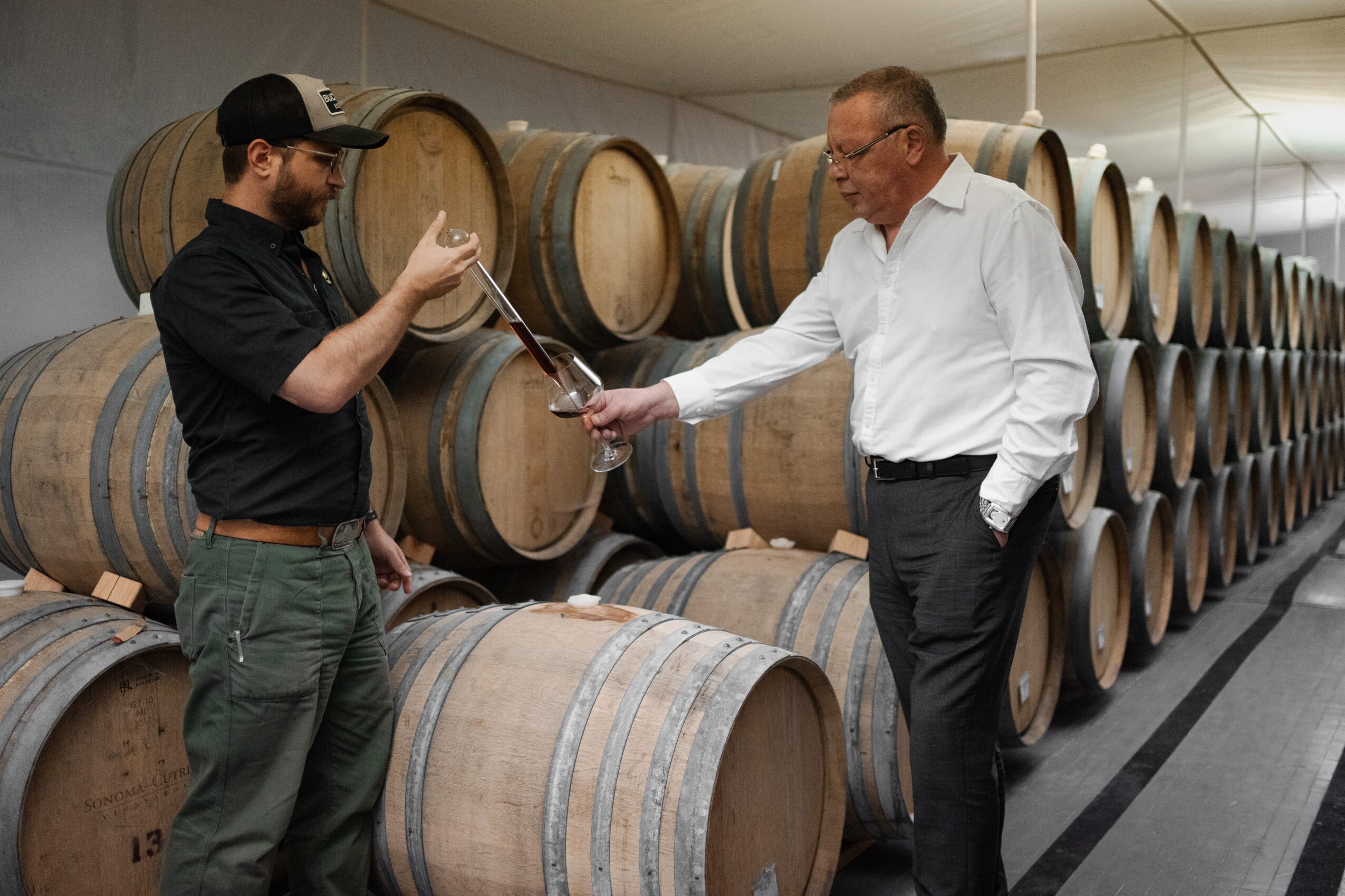 Two men in a wine cellar exchanging a bottle, with stacked wine barrels in the background.