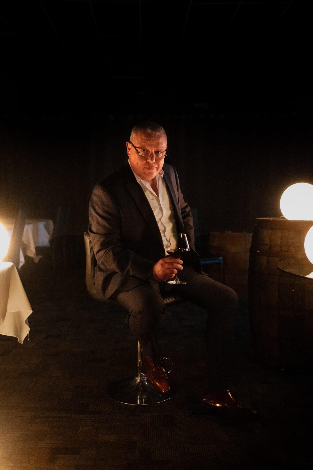Man in suit sitting on a stool, illuminated by a round light, in a dark setting.