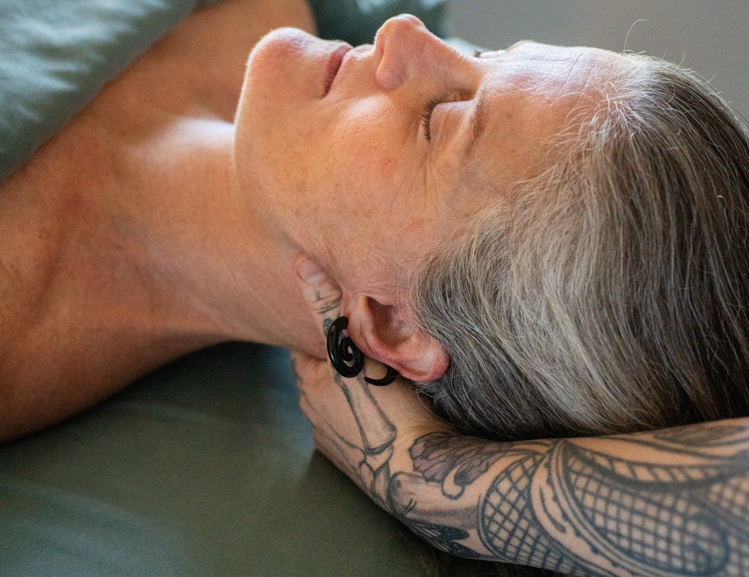 Woman with gray hair lying on her side with head resting on a tattooed arm, eyes closed, peaceful expression.