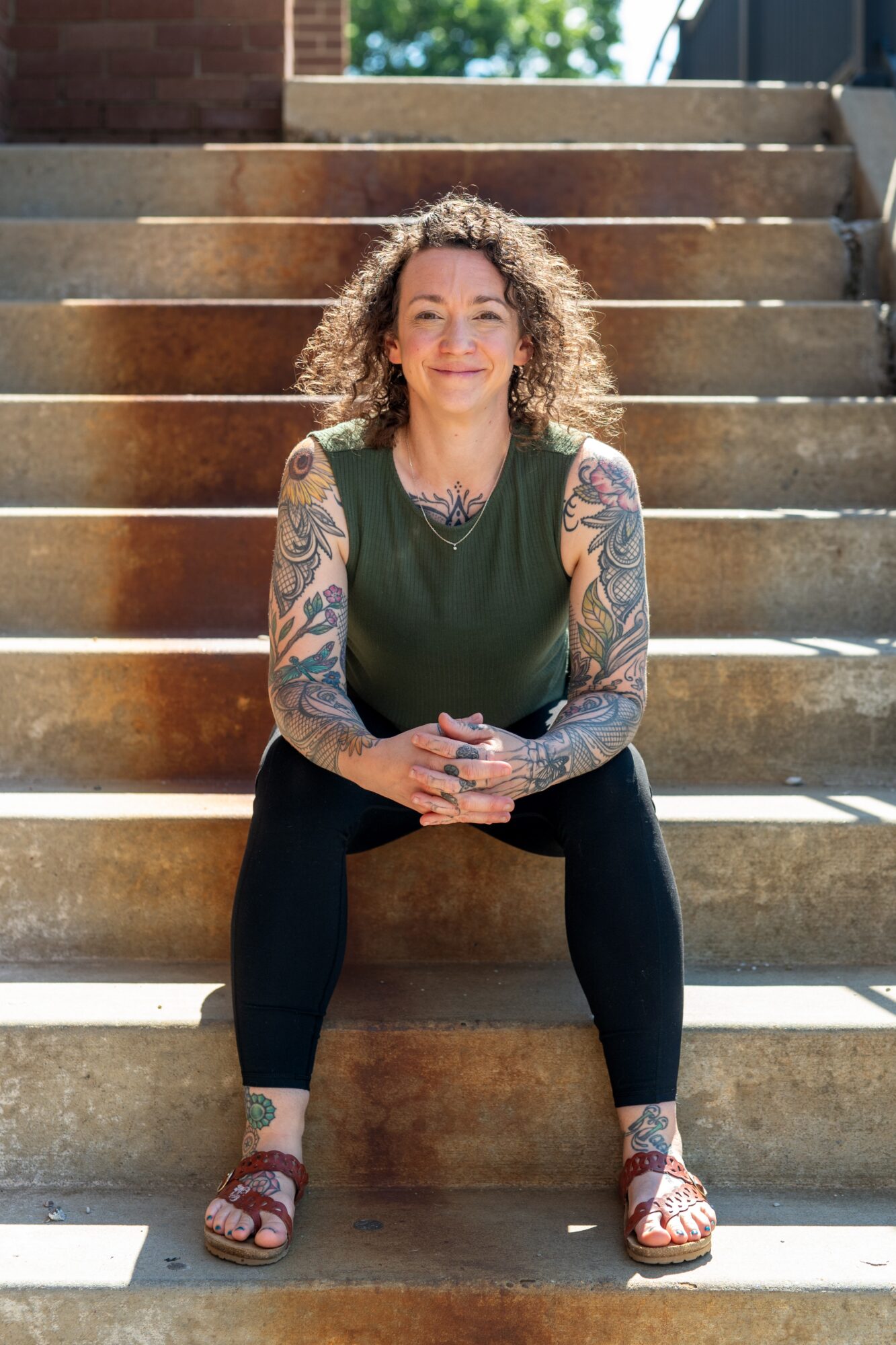 Woman with curly hair, tattoos on arms, sitting on stairs outdoors, smiling, wearing a green top, black pants, sandals.