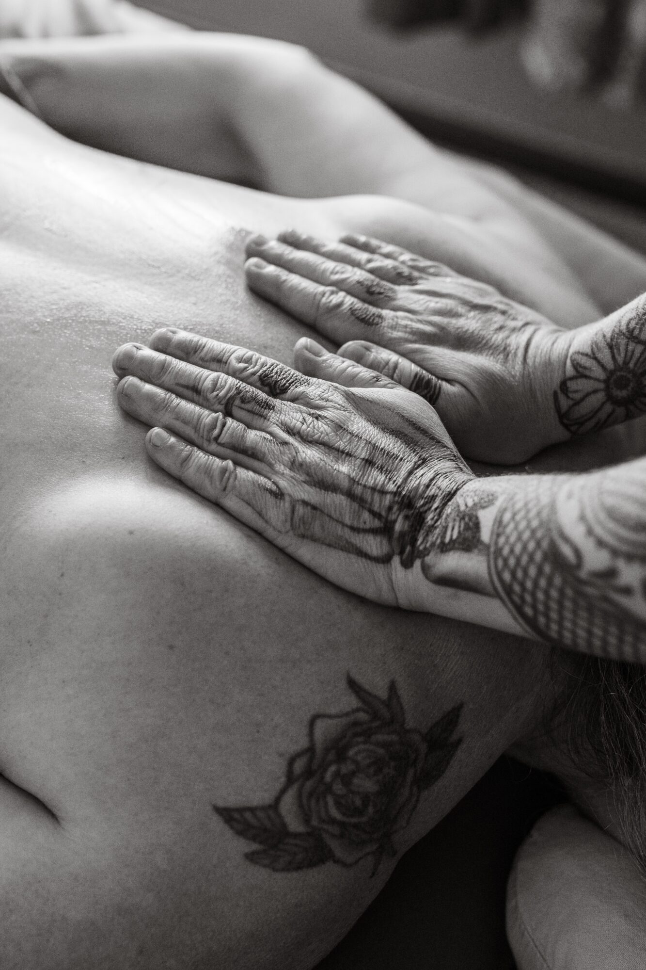 Two hands with tattoos rest on a person's bare chest, which has a floral tattoo. The image is in black and white.