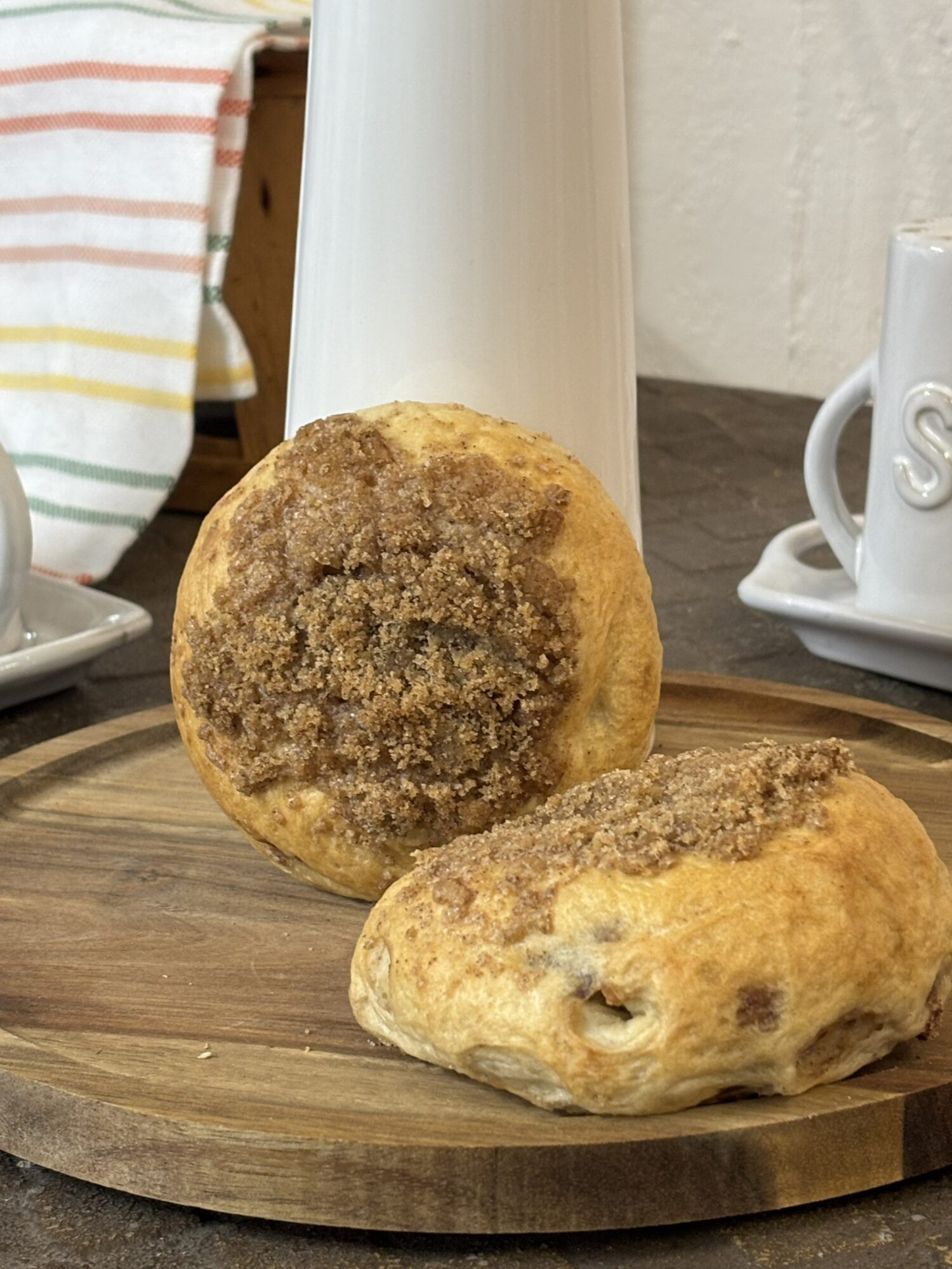 Two fried pastries on a round wooden board, with a white pitcher and cups in the background.