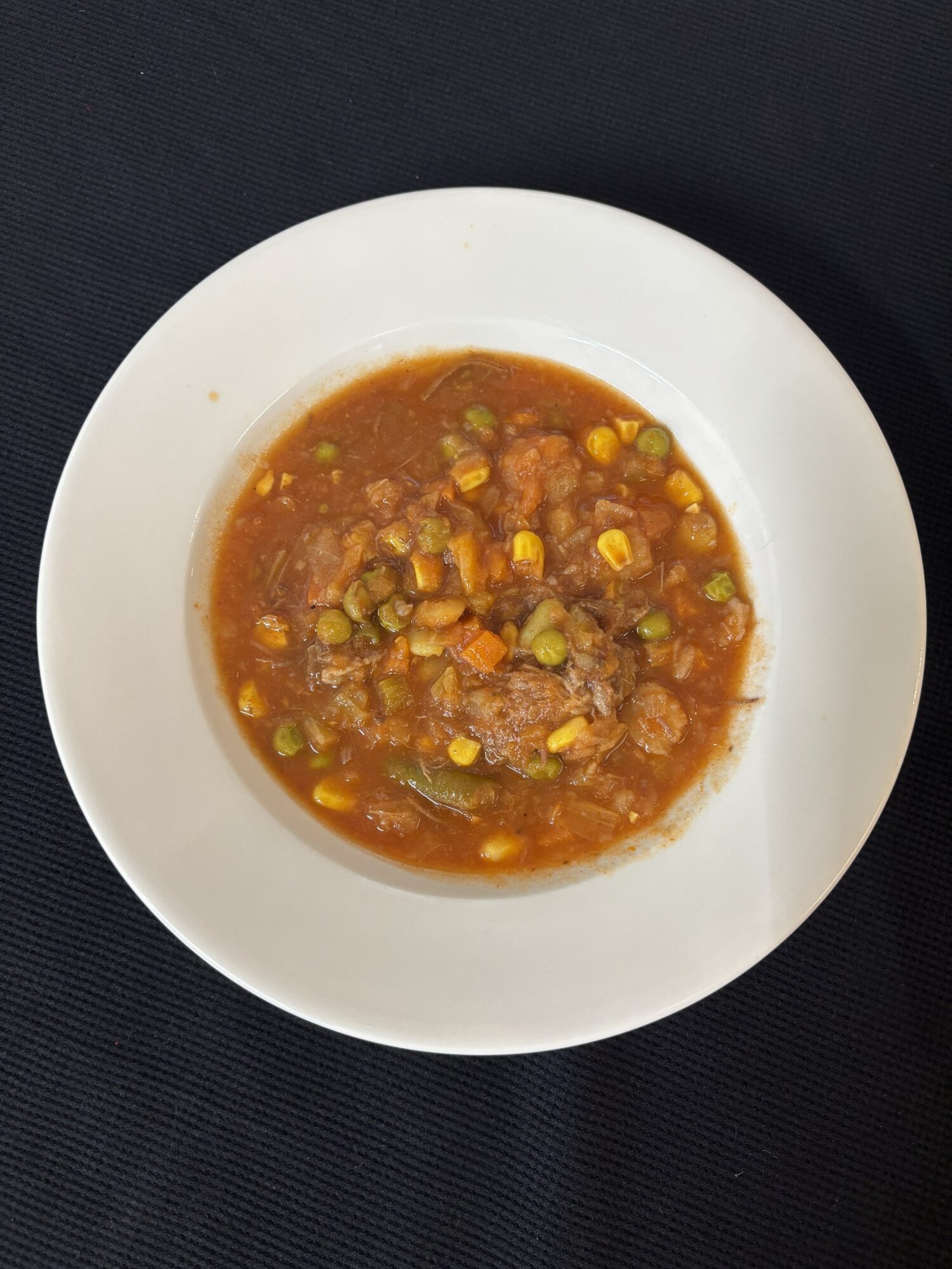 Bowl of vegetable soup with corn, peas, and carrots in a broth on a white plate, black background.