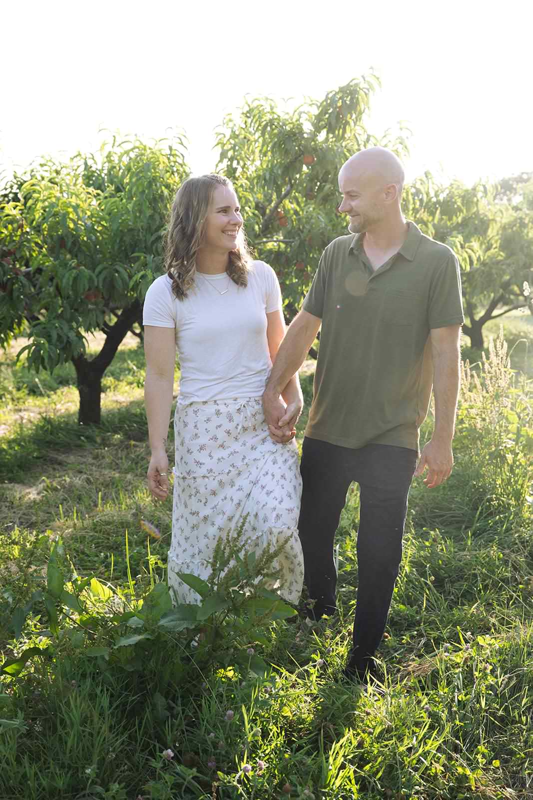 A couple holding hands walking outdoors in a grassy area with trees, smiling at each other.