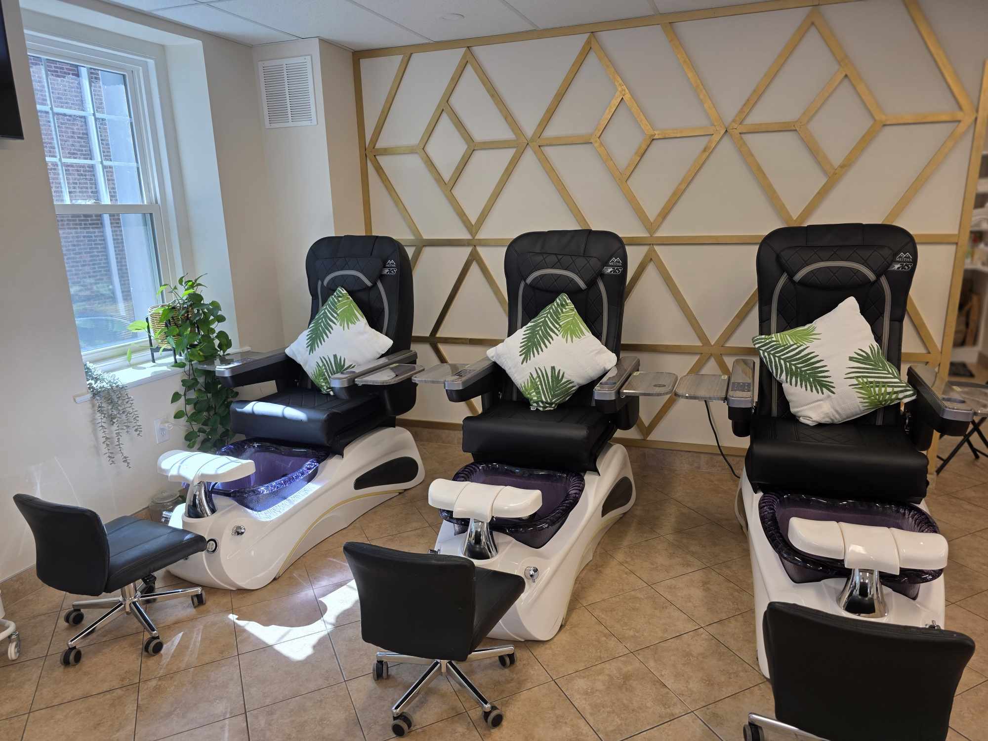 Three pedicure chairs with black cushions and white bases, each with a pillow, in a room with a geometric wall design and window.