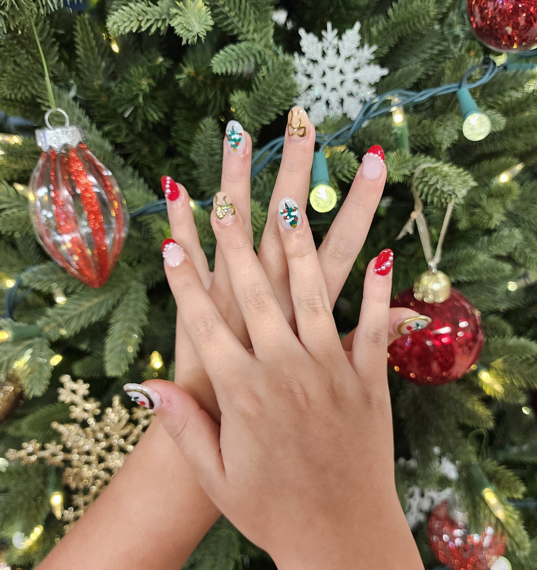 Two hands with decorated nails in front of a Christmas tree with ornaments and lights.