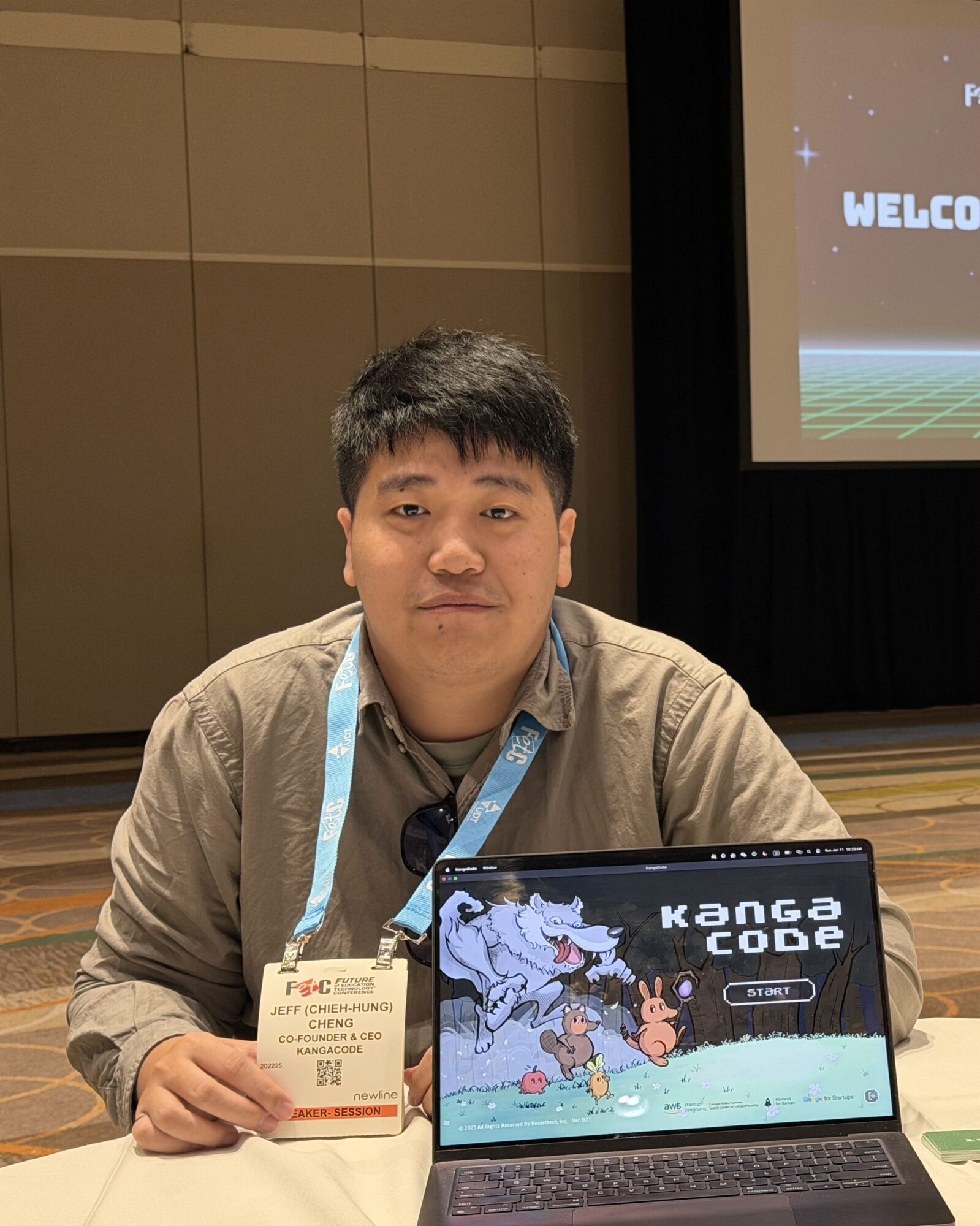 Young man sitting at a table holding a laptop with a game titled 'KONG CODE' on the screen, in a conference room.