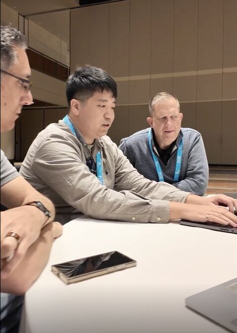 Three men sitting at a table, focused on a laptop, in a conference room with beige walls.
