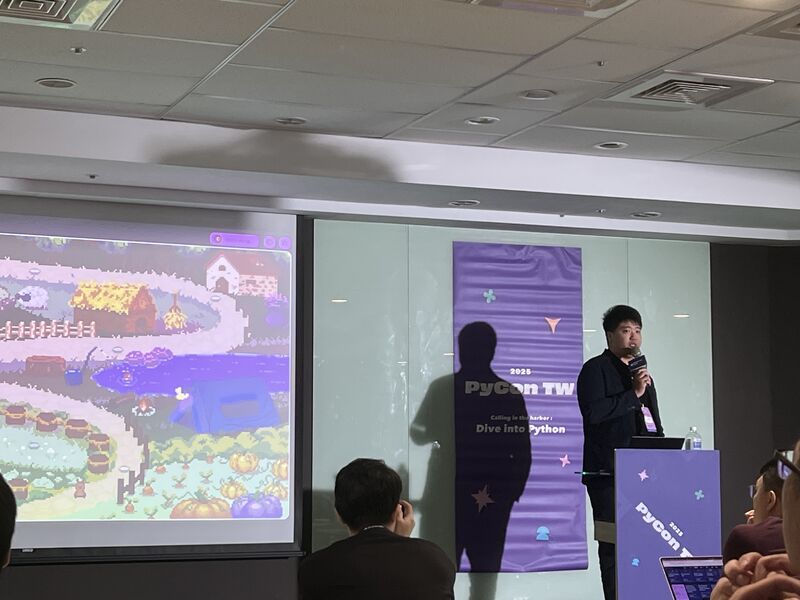Person standing at a podium giving a presentation in a conference room, with a large screen displaying a colorful pixel art scene.