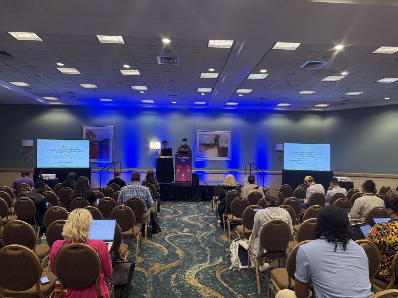 Conference room with audience facing stage, two screens displaying presentation slides, and two speakers at podiums illuminated by blue lighting.