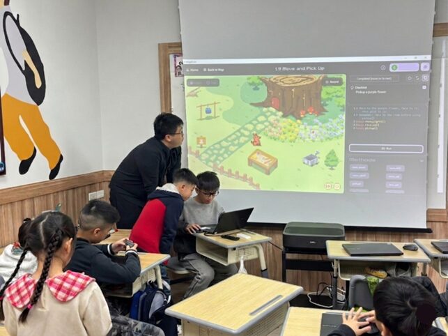 Teacher and students in classroom watching a projected screen with a game or educational content, desks and chairs visible.