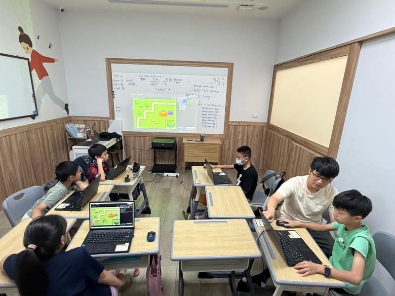 Classroom with students at desks using laptops, teacher at whiteboard, and a mural on the wall.