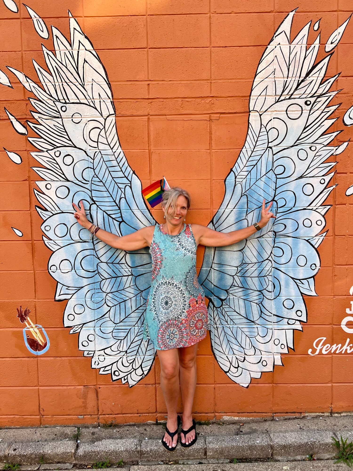 Person with large butterfly wings painted on a wall, arms outstretched, standing on a sidewalk, brick wall background.