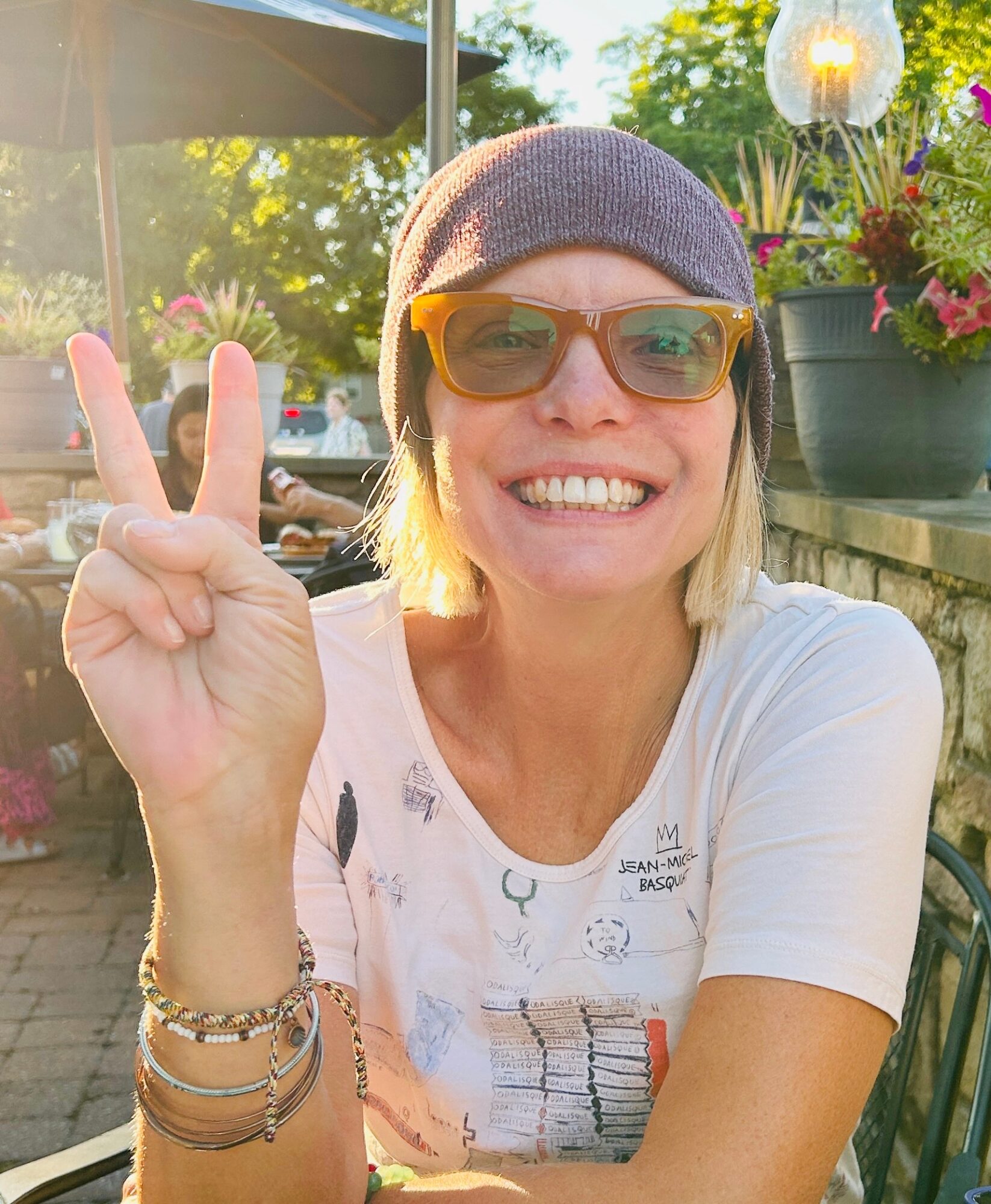 Woman smiling outdoors, wearing sunglasses and a beanie, making a peace sign with her right hand, sunlight in background.
