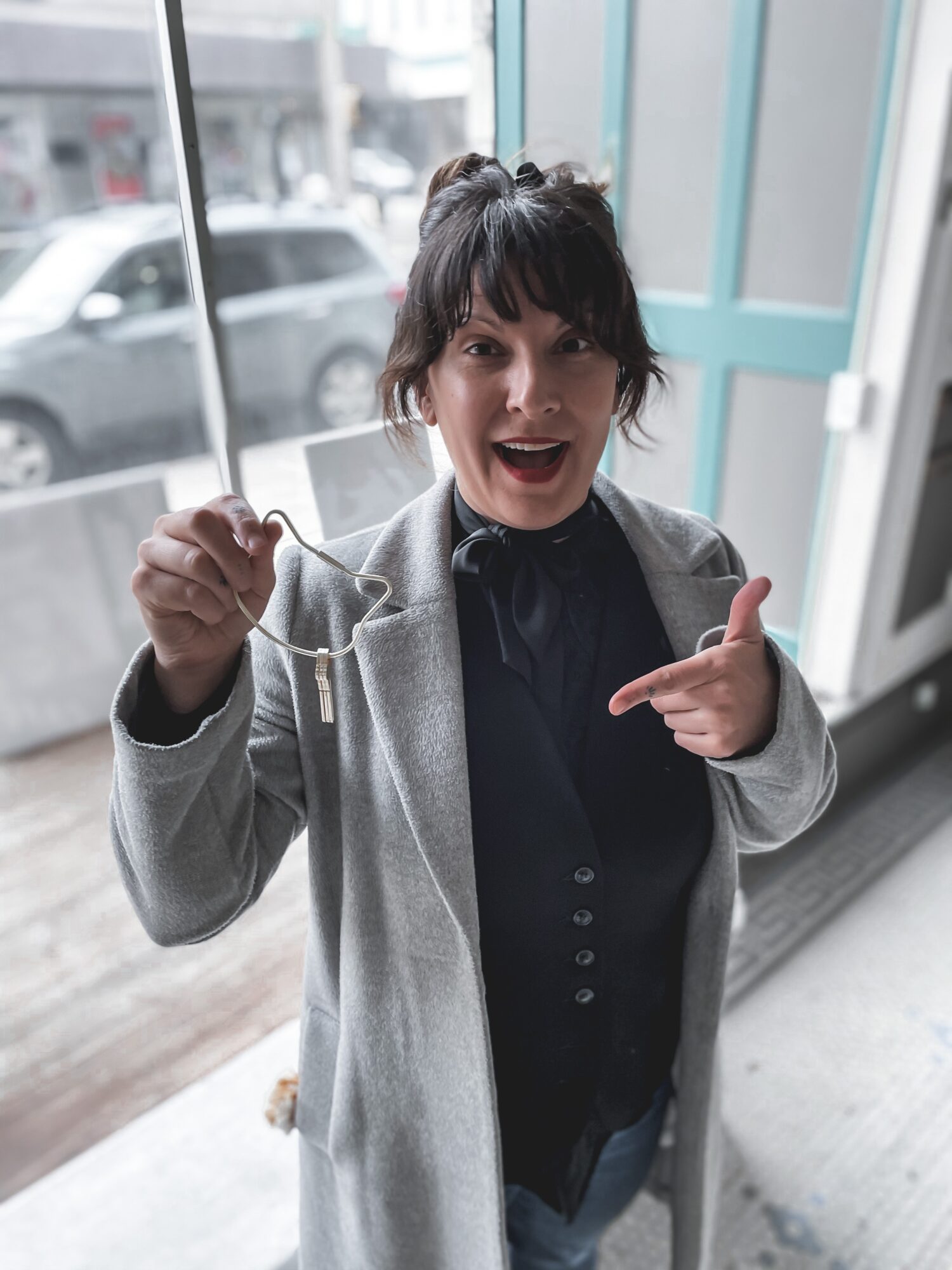Woman with short dark hair smiling, pointing at herself, holding a string, standing indoors near large windows.