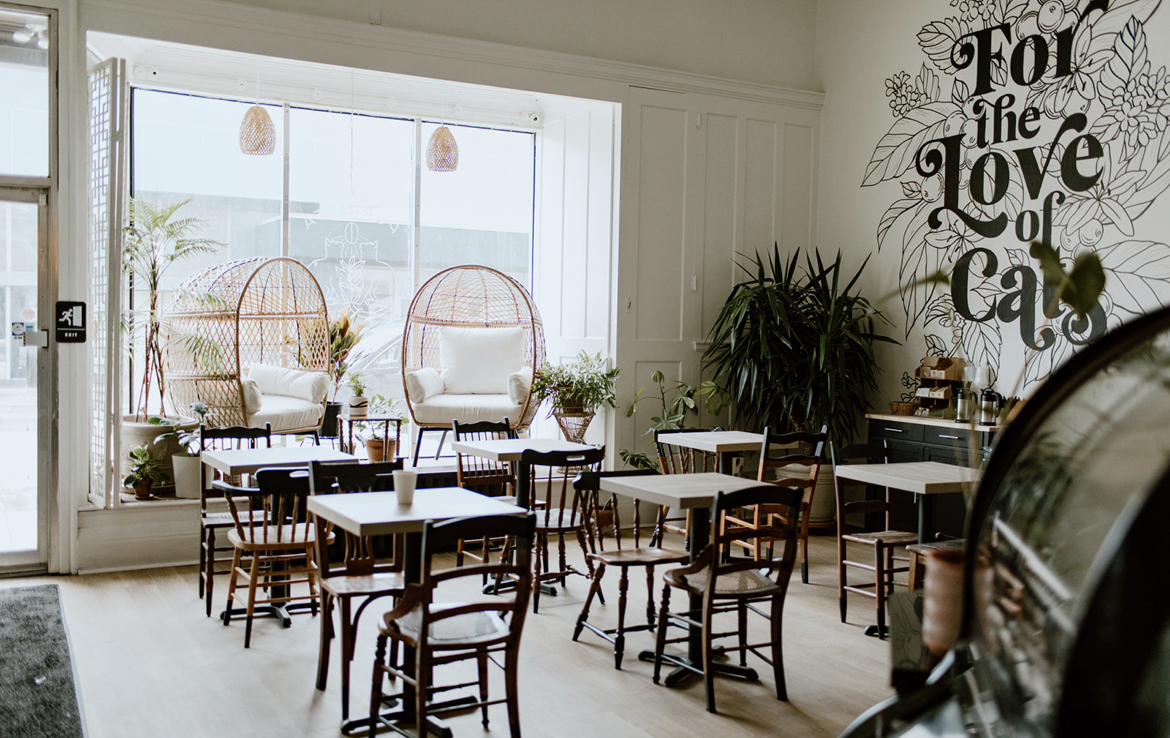 Interior of a cafe with tables and chairs, large window, and wall art reading 'For the Love of Coffee'.
