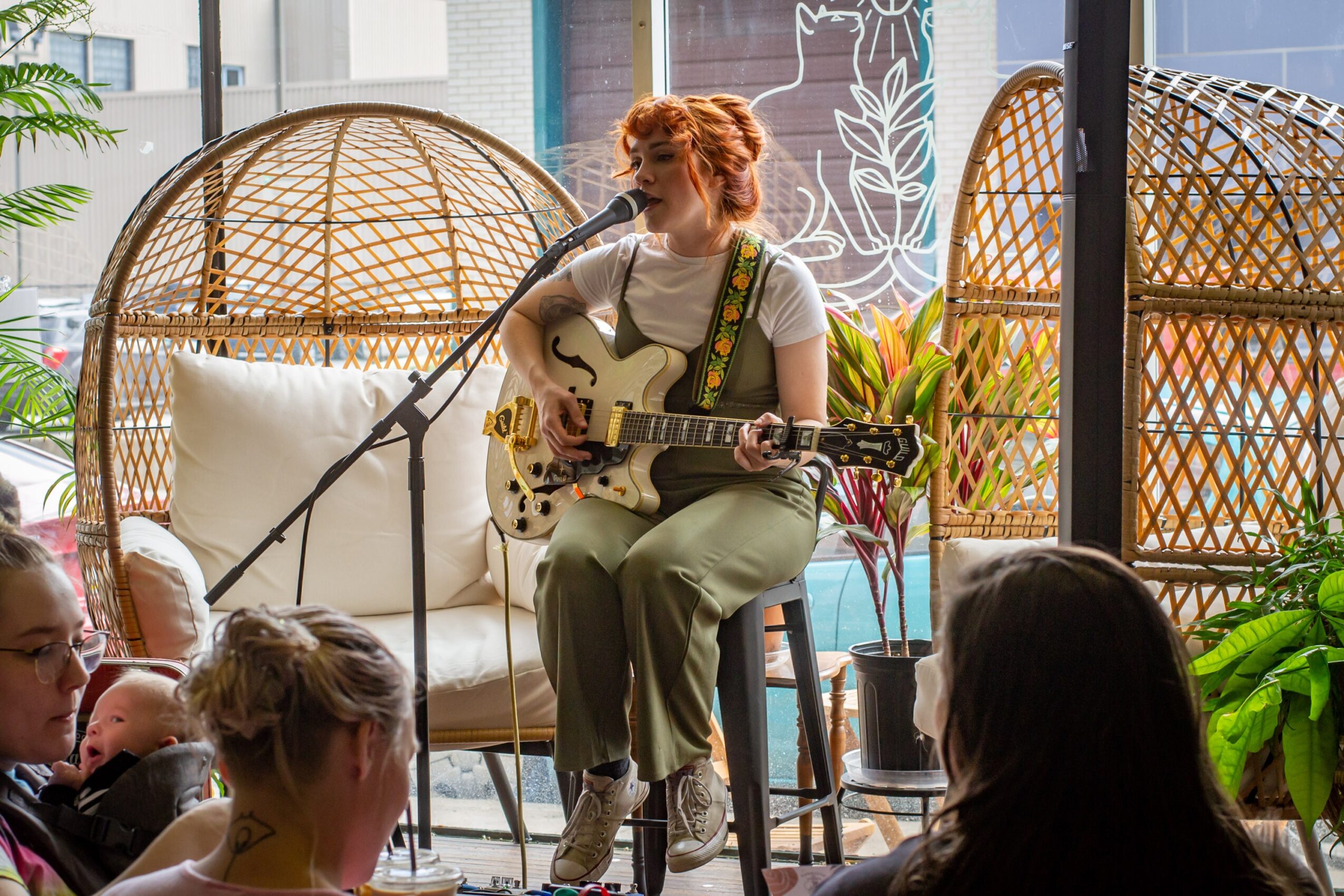 A woman with red hair playing an electric guitar on a stool, surrounded by wicker chairs and plants in a cozy indoor space.
