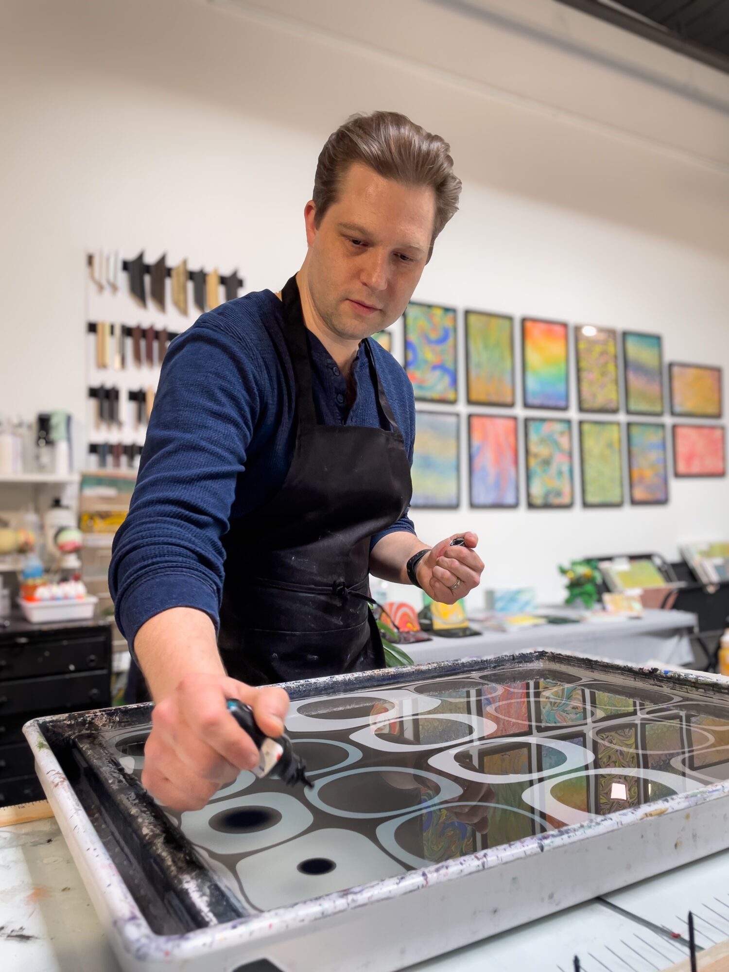 Man working on glass art project in studio with colorful paintings on wall behind him.