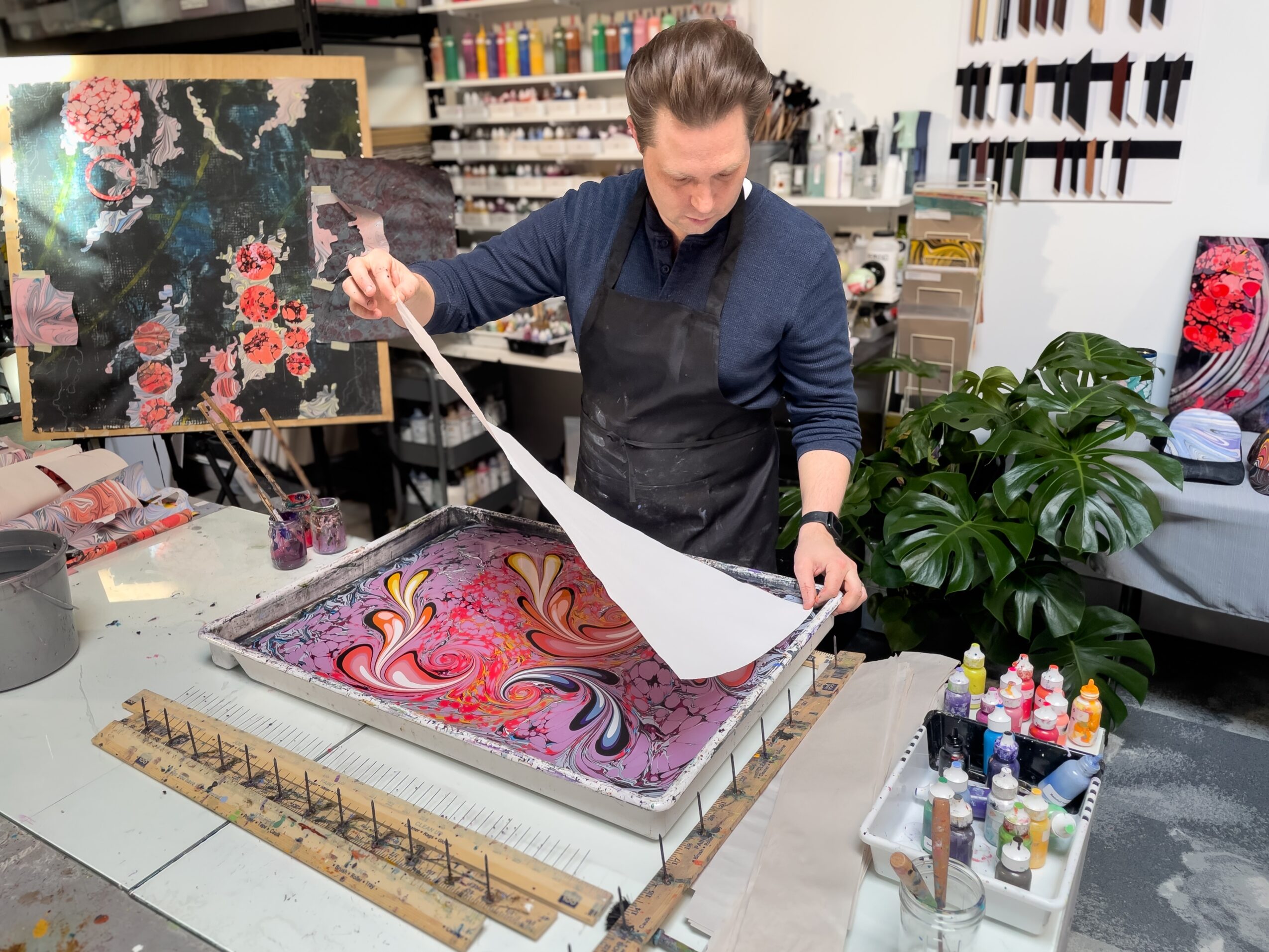 Person working on a colorful marbled paper in an art studio, surrounded by art supplies and plants.