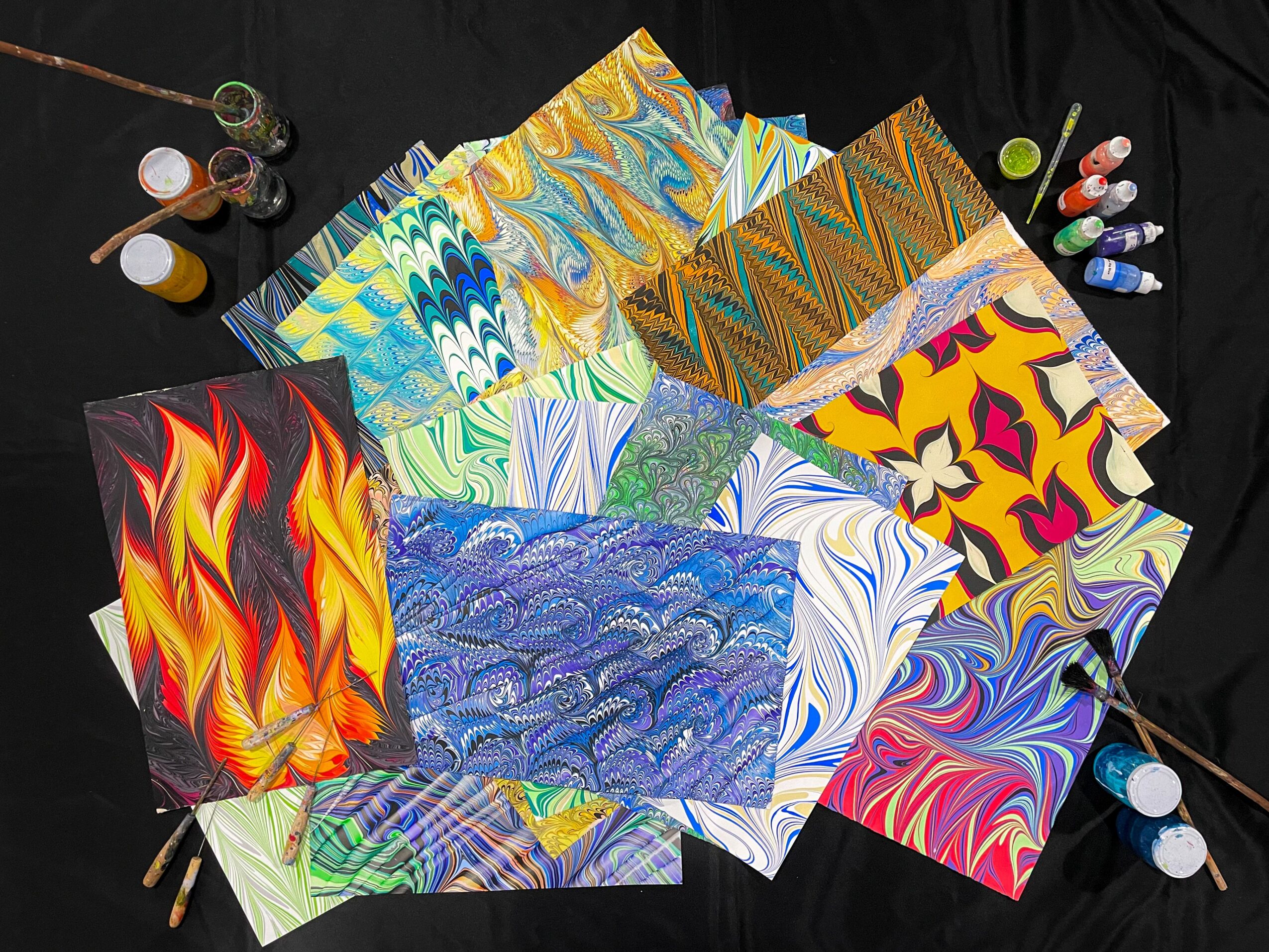 Colorful marbled paper sheets arranged in a fan shape with bottles of paint and brushes around them.
