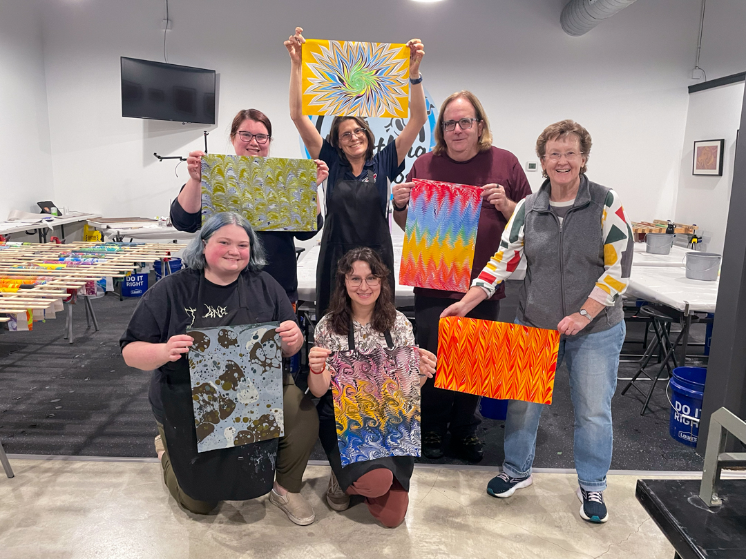 Group of seven women holding colorful fabric pieces in a room with tables and a TV screen.