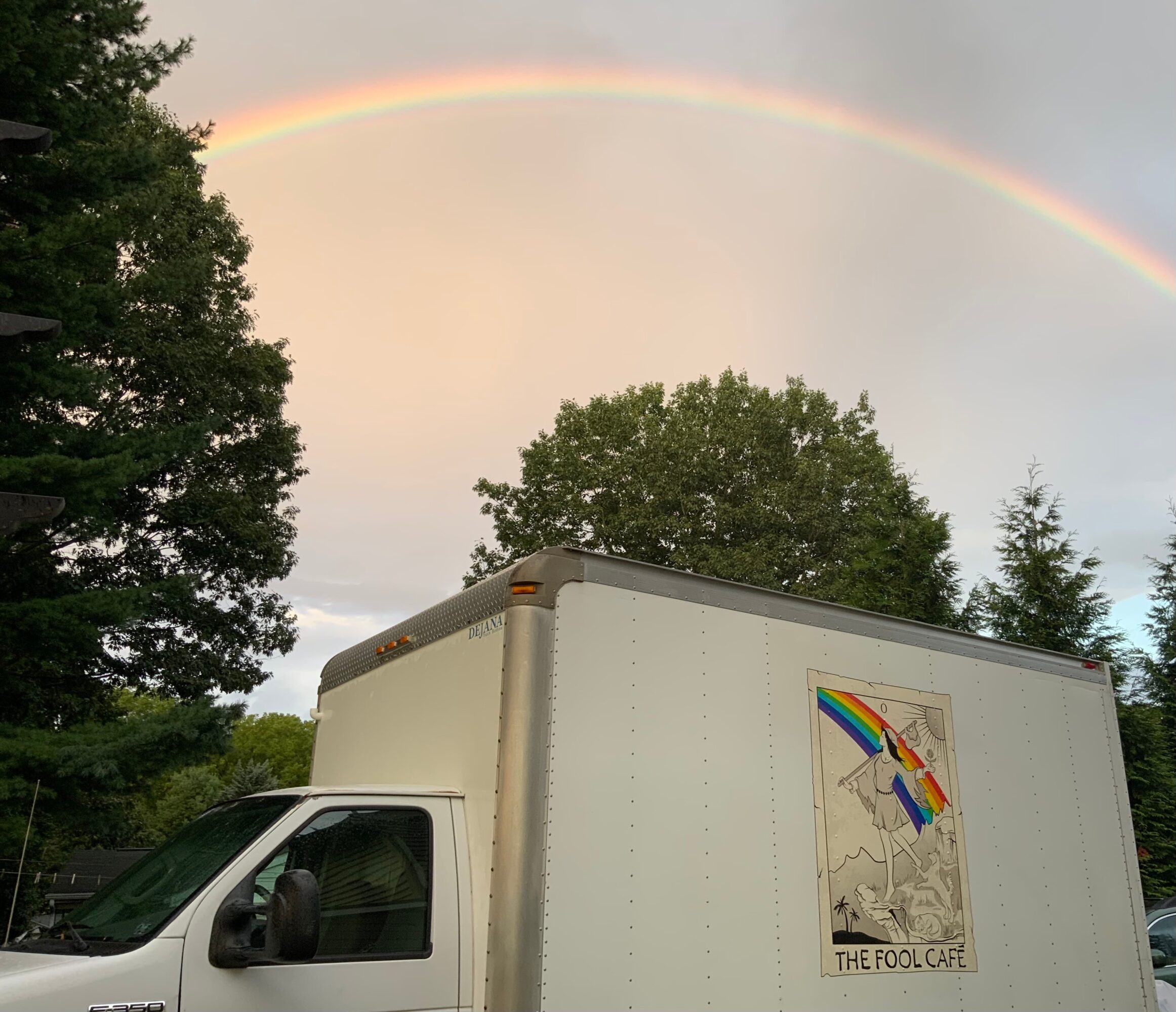 Rainbow arching across the sky above trees and a truck with a colorful logo, partly cloudy sky, and sunlight.