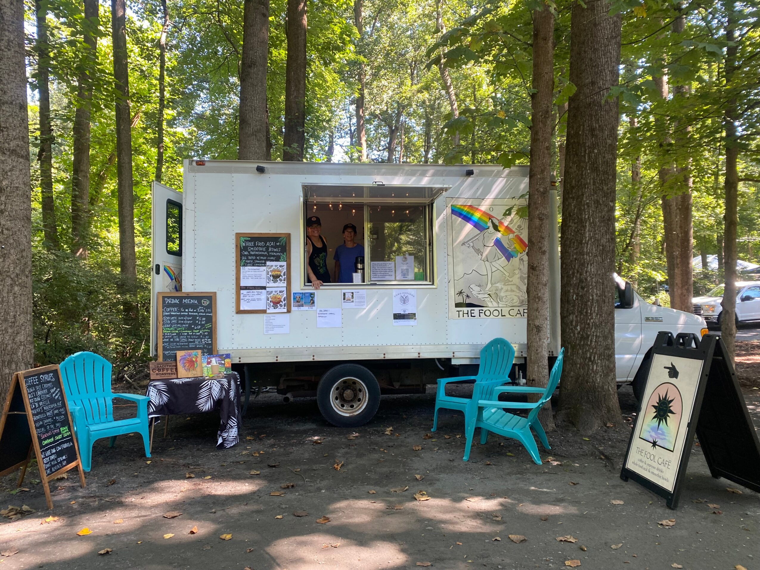 Food truck with people inside, surrounded by trees, with colorful chairs and a chalkboard outside, in a forest setting.