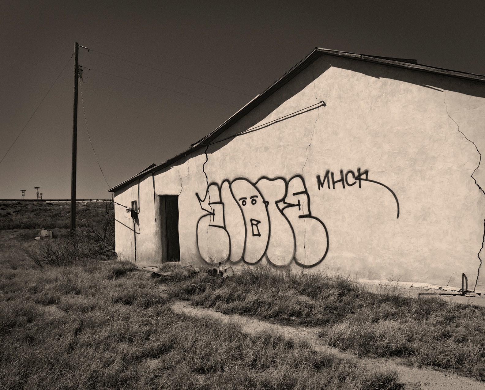 A building with graffiti on its wall, situated in a rural area with a grassy foreground and a utility pole in the background.