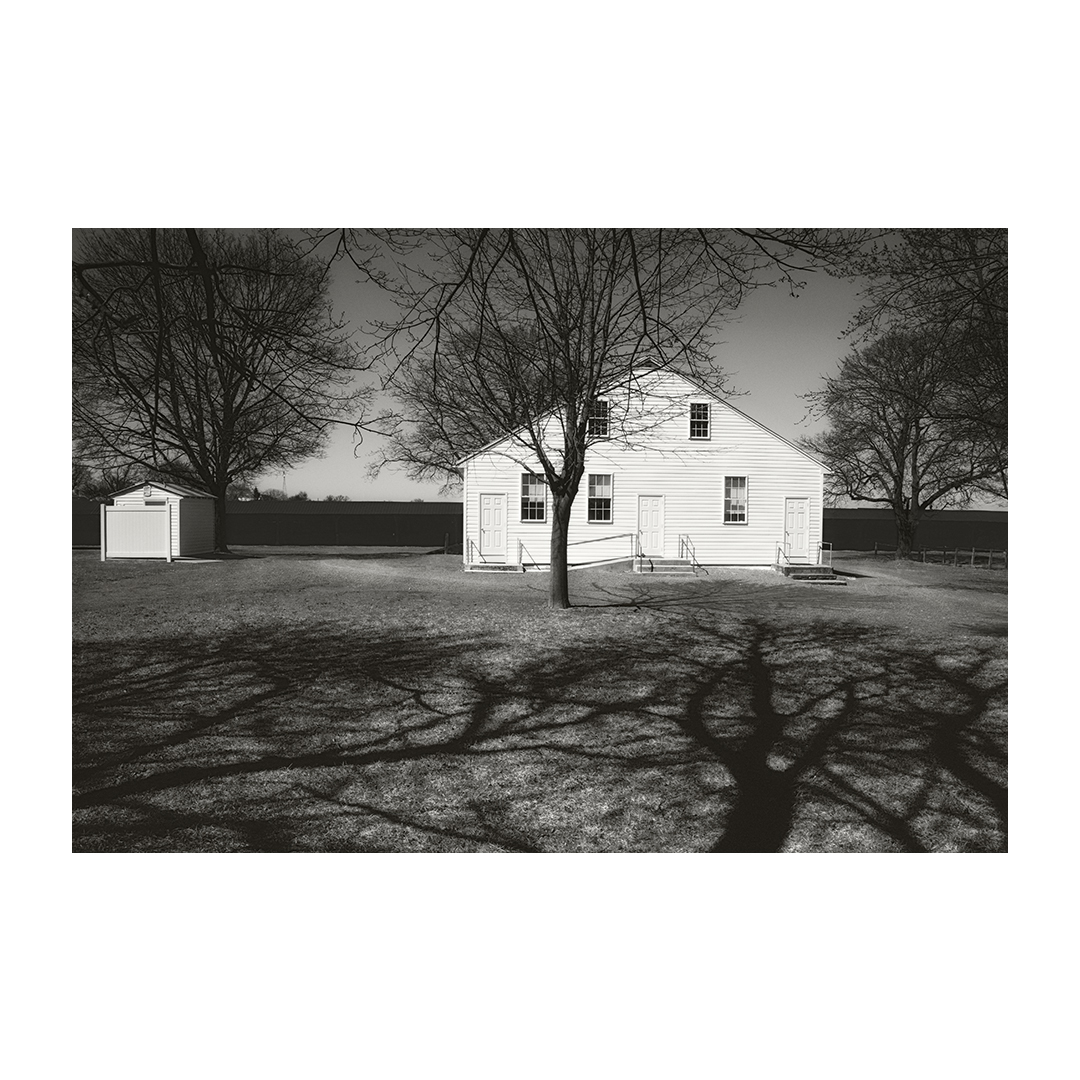 A white house with trees in front, shadows on the ground, and a small shed to the left, in black and white.