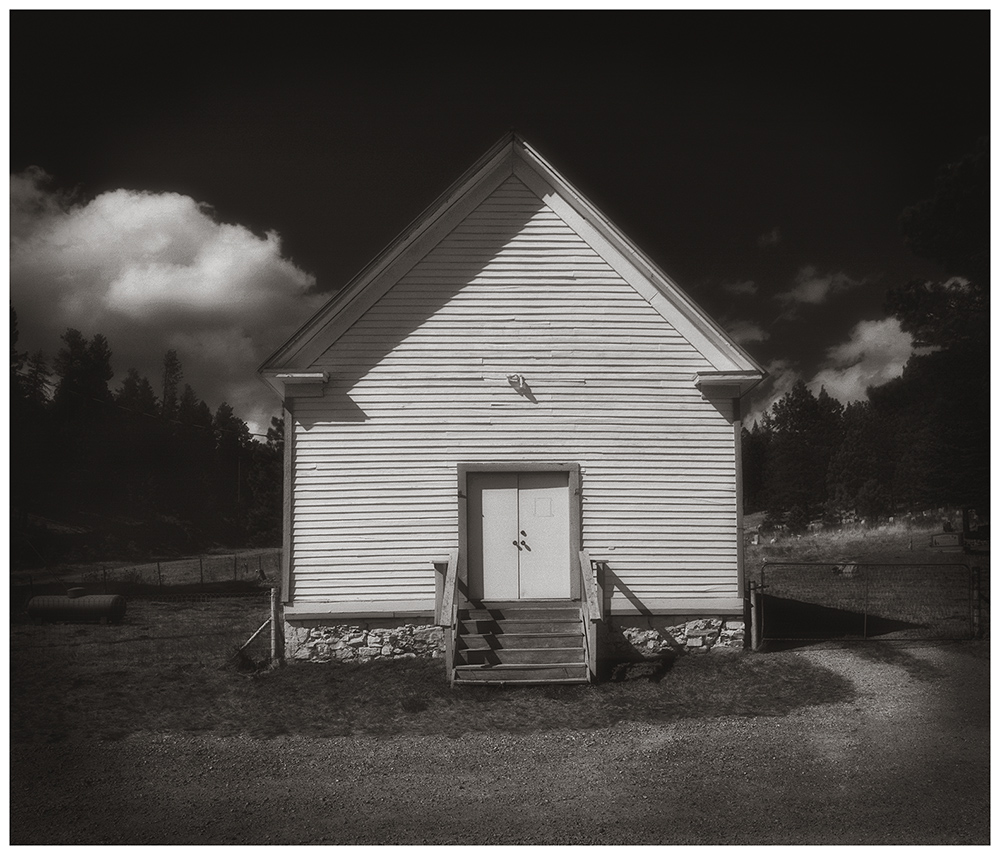 Small white building with stairs and a door, set against a cloudy sky and trees in the background.