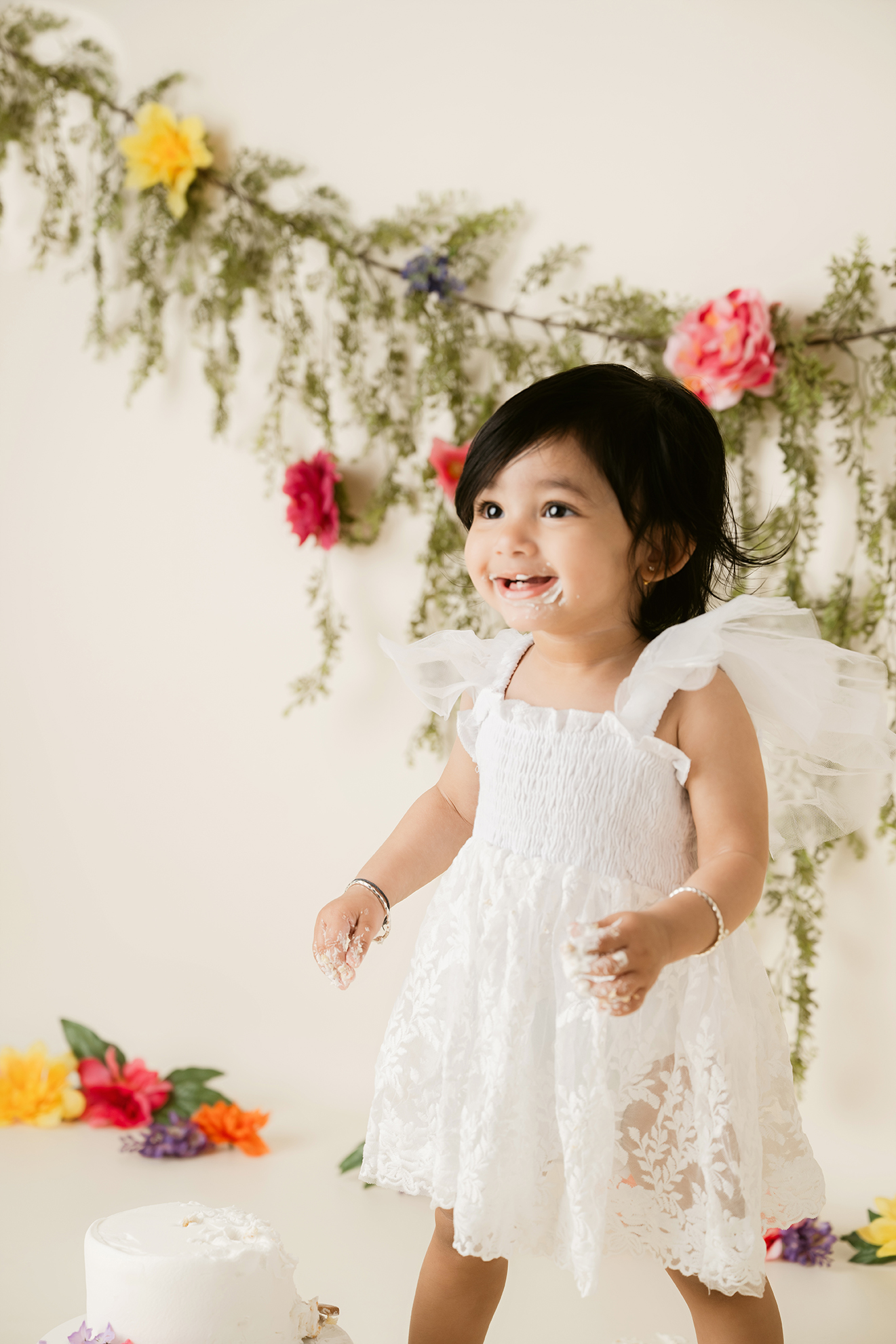 Young girl in a white dress smiling, standing in front of a floral backdrop with colorful flowers and greenery.