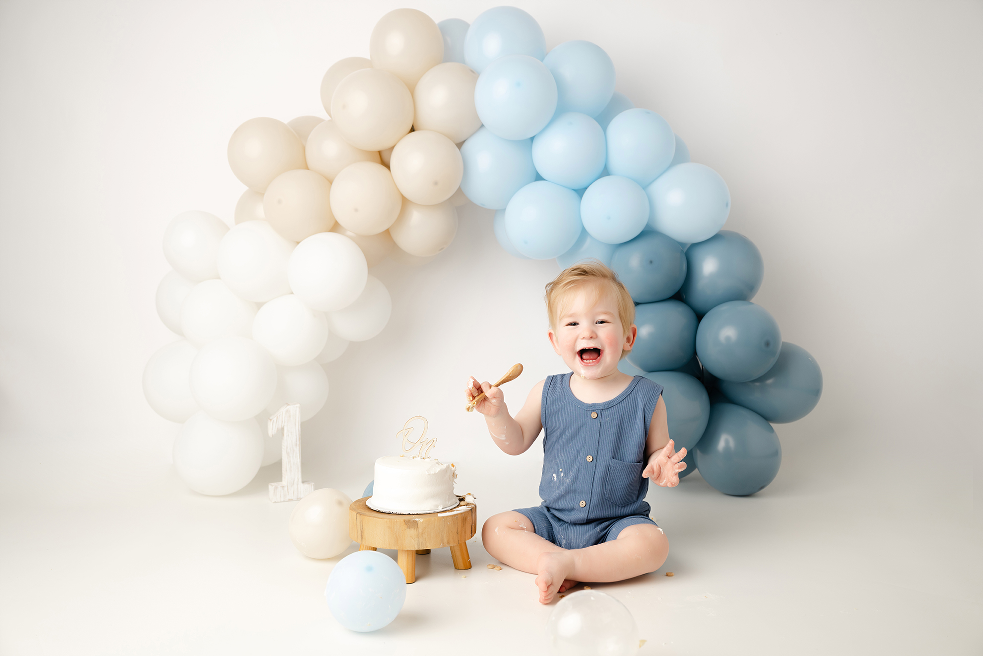 A laughing child sitting on the floor in front of a balloon arch with white, beige, and blue balloons, celebrating a first birthday.