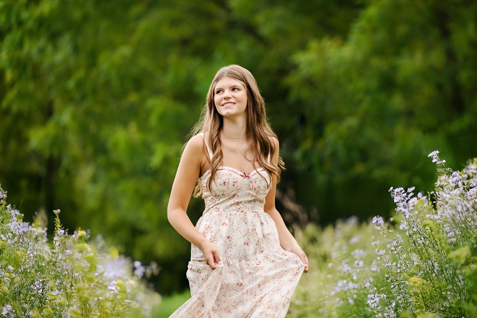 Young woman in a floral dress standing outdoors among tall flowers, smiling and looking upward, with green trees in background.