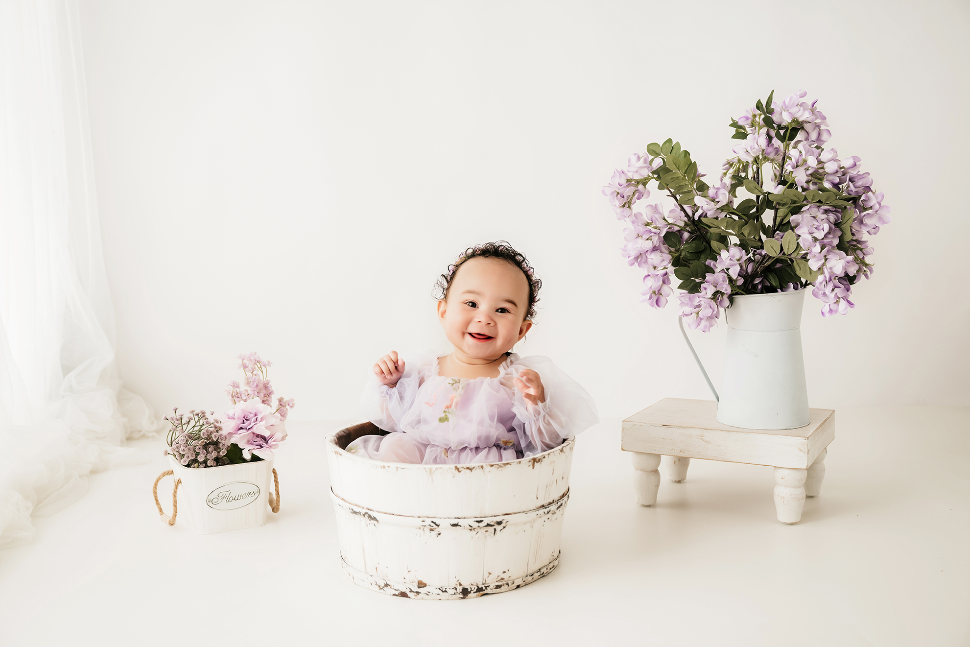 Child smiling in a white bucket with flowers, on a white background, with a small table and flower pot nearby.