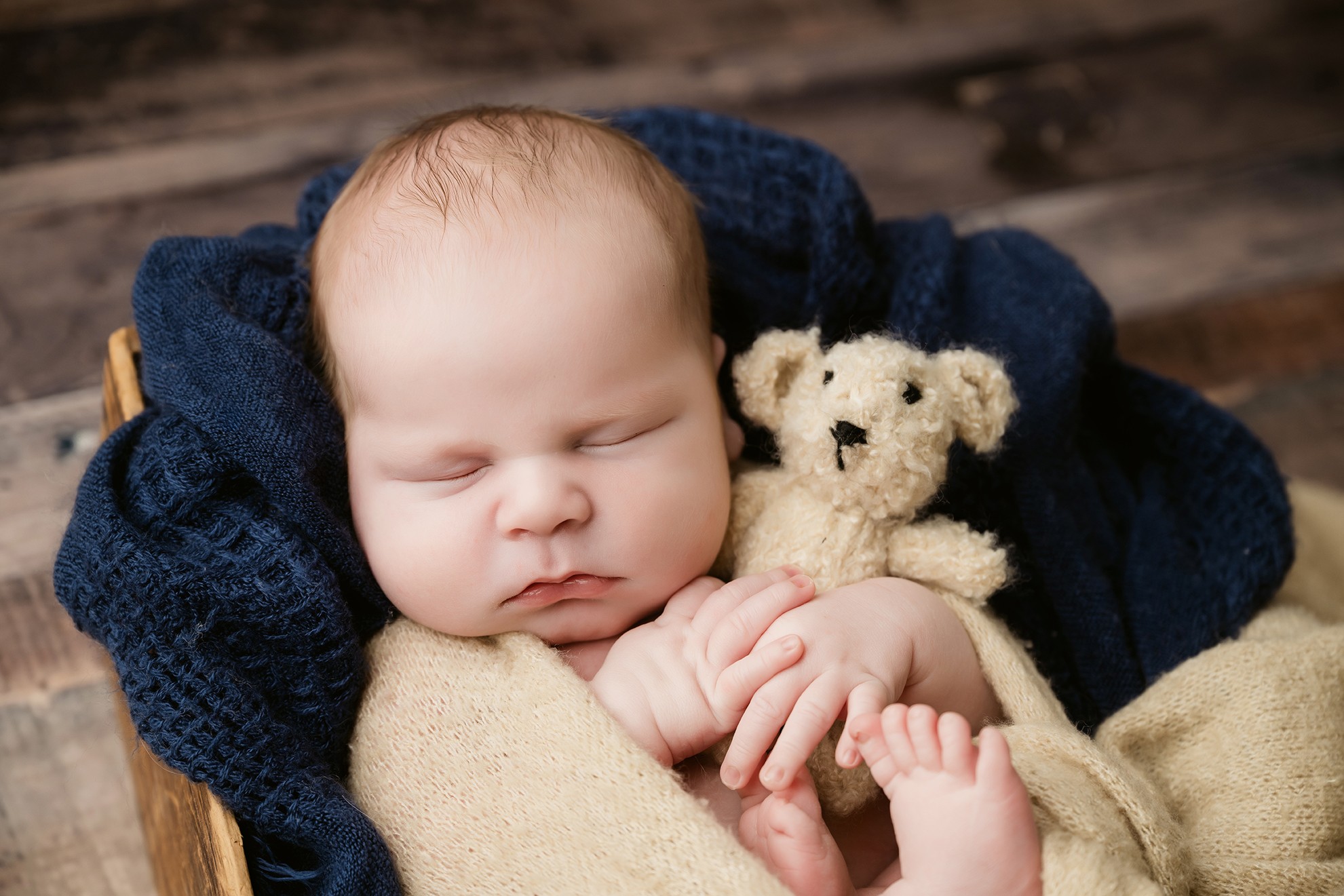 Baby sleeping with a teddy bear, wrapped in a blanket, on a dark wooden surface.