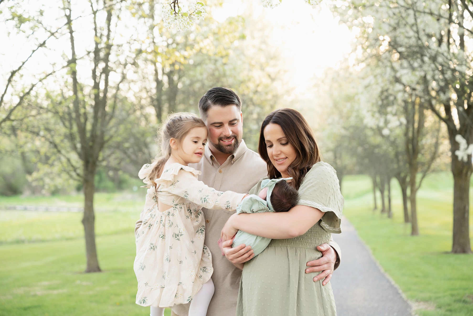 Family of four outdoors, woman holding a baby, man and girl looking at the baby, trees in background