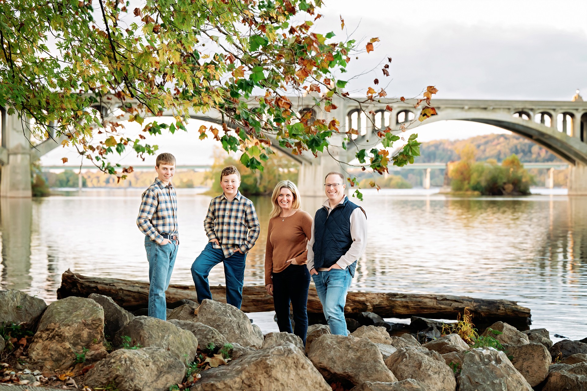 Four people stand on rocks by a river with a bridge and trees in the background.