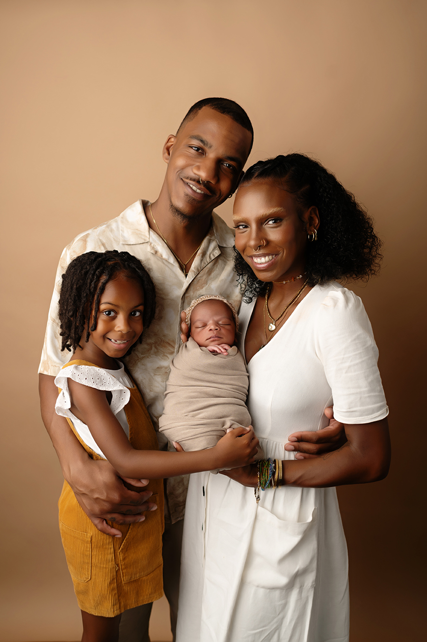 Family of four with a man, woman, young girl, and newborn baby, smiling together against a plain background.