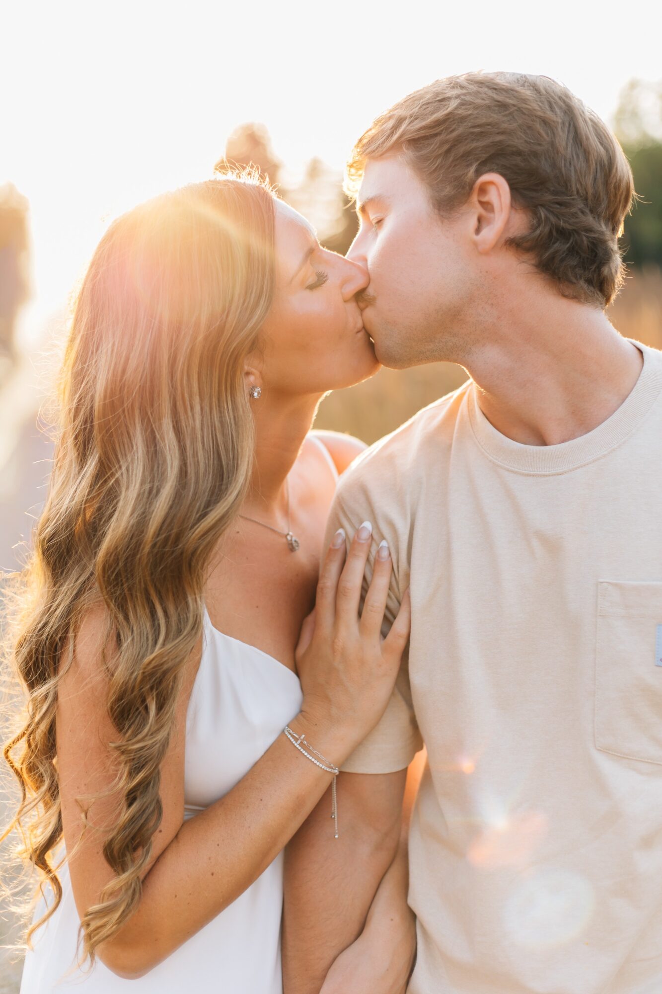 Couple kissing outdoors with sunlight, woman has long wavy hair, man has short hair, woman wears a white top, man wears a light-colored shirt.
