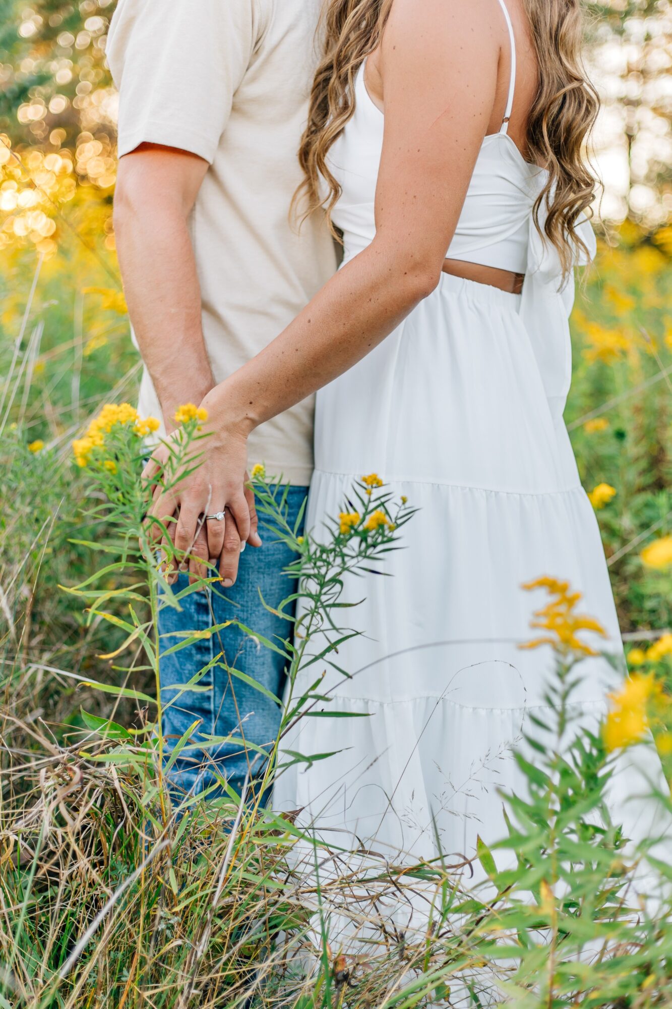 Two people holding hands in a field of yellow flowers, one wearing a white dress and the other in a light-colored shirt and jeans.