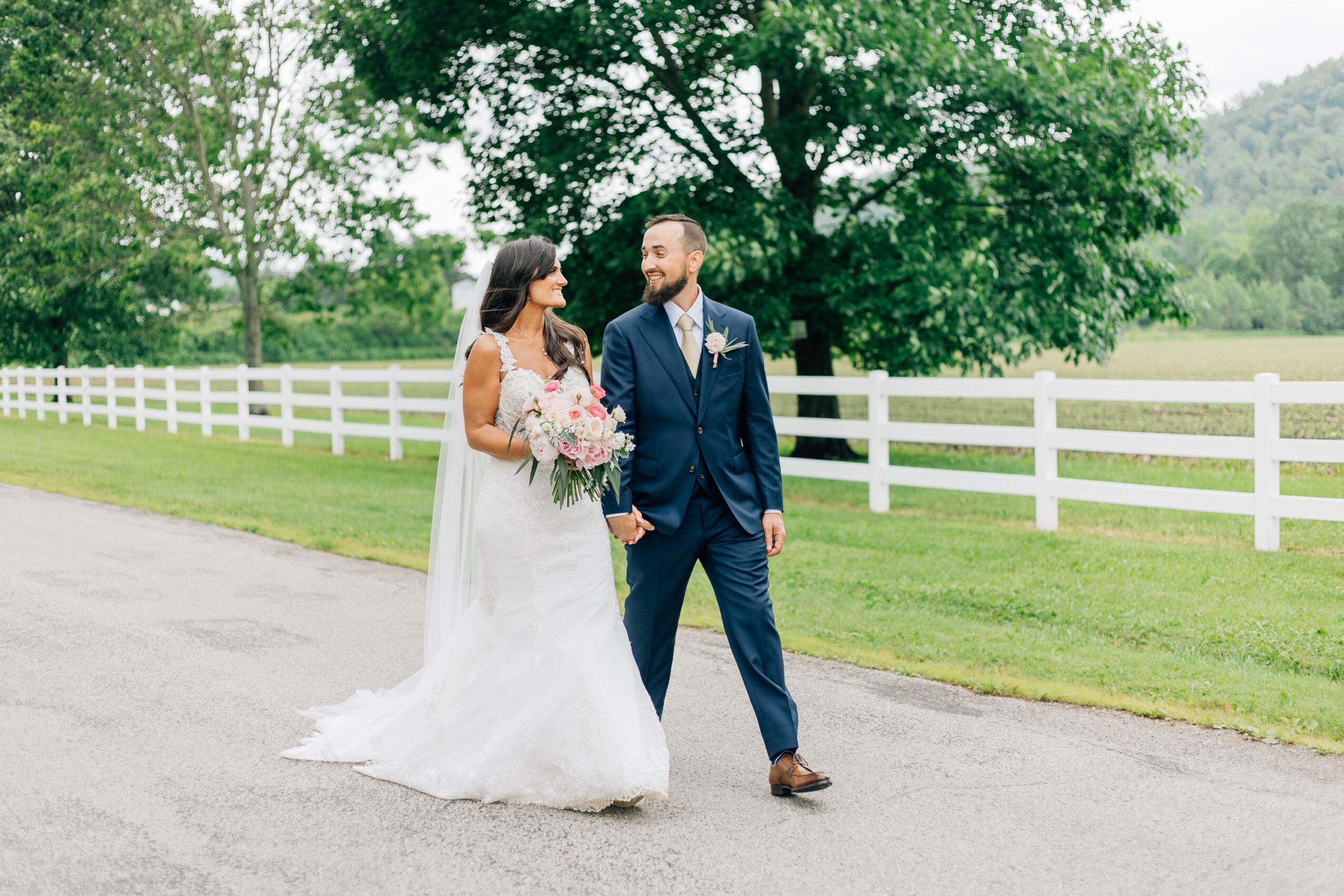 Bride and groom walking outdoors on a paved path, holding hands, with a white fence and green trees in the background.