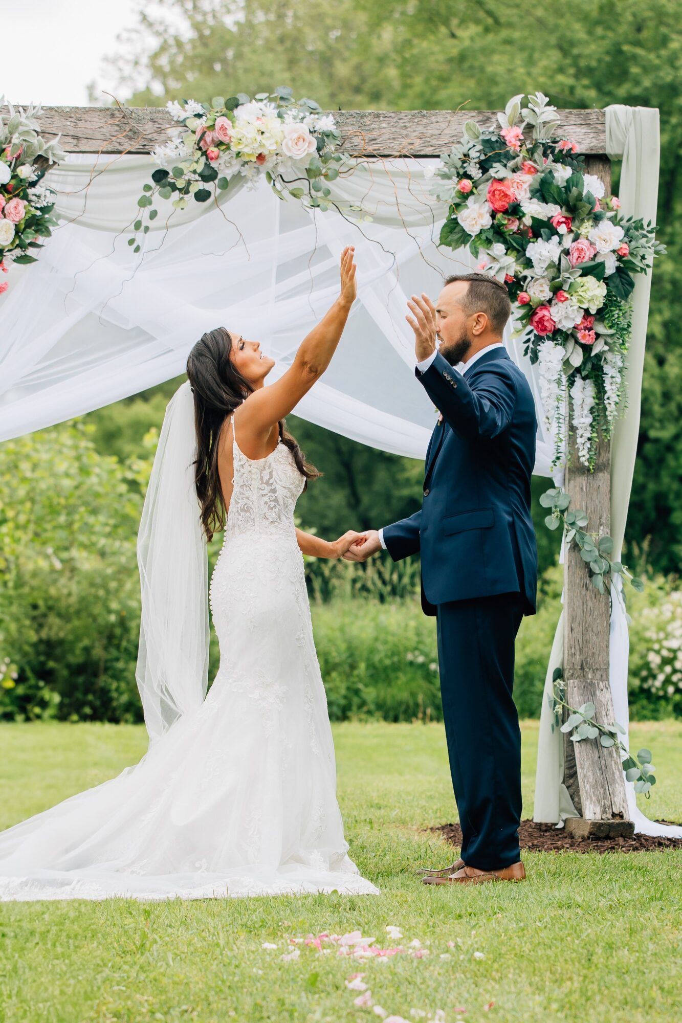 Bride and groom holding hands and smiling under a decorated wedding arch outdoors, surrounded by greenery