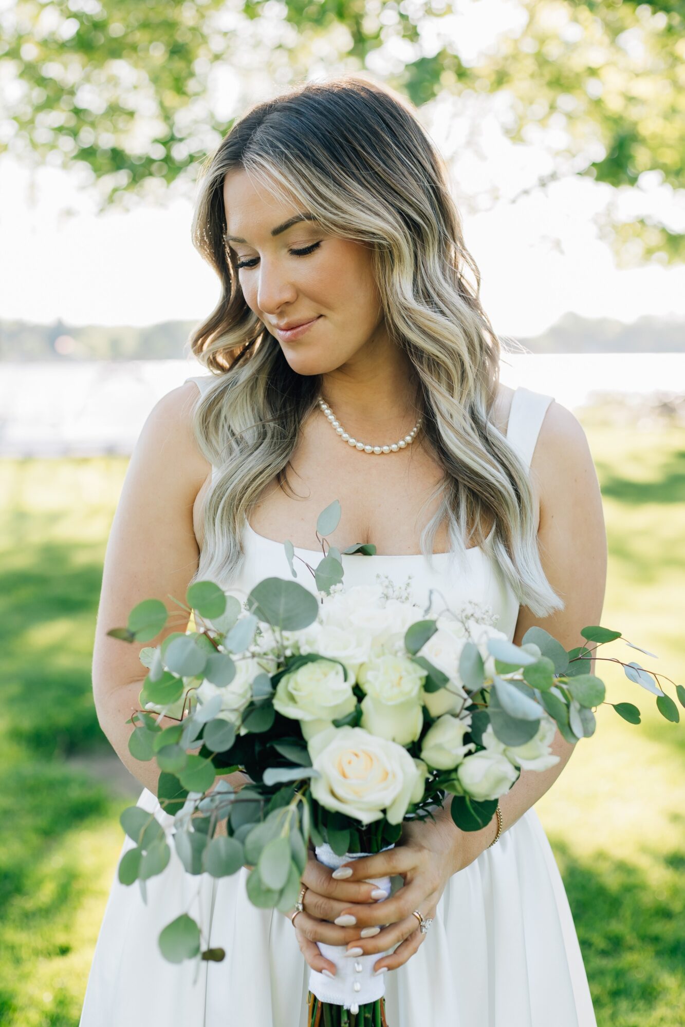Woman in white dress holding a bouquet of white roses and greenery outdoors, smiling with eyes closed.