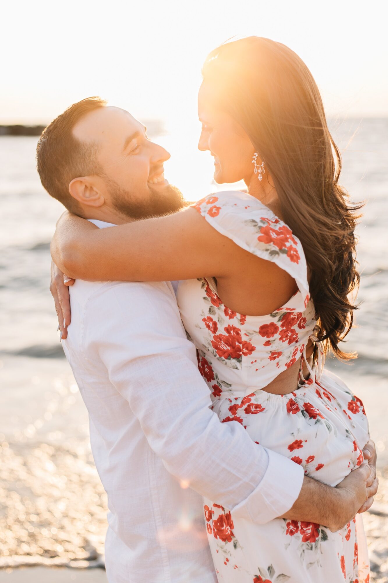 A couple embraces on a beach at sunset, smiling at each other, with the ocean in the background.