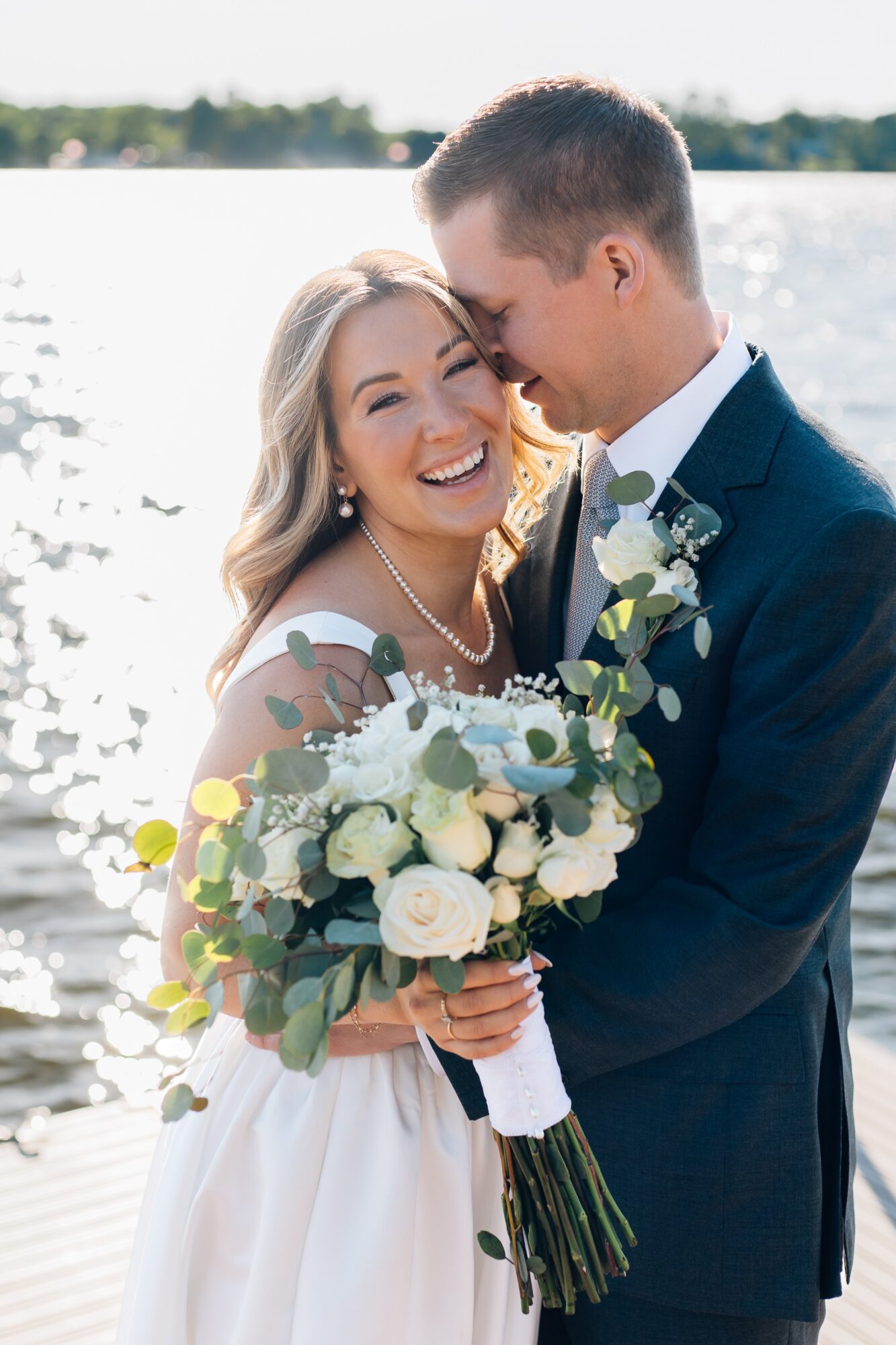 Bride and groom smiling, holding a bouquet, near a body of water, outdoors, sunny day.