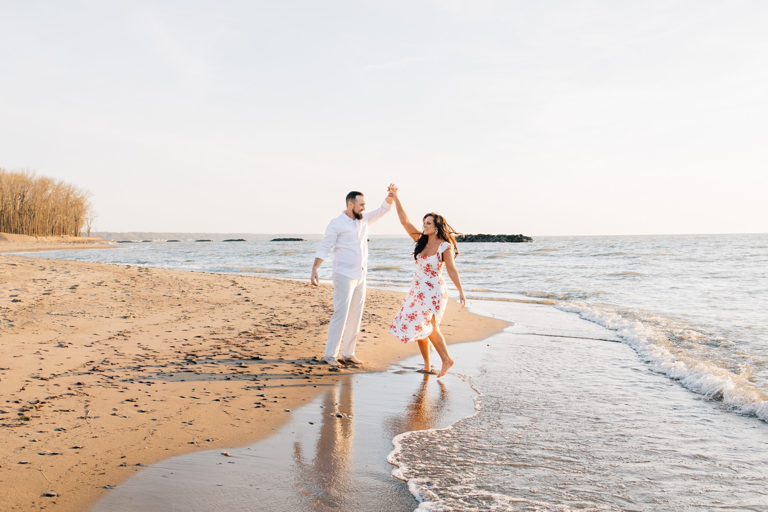 A couple dancing on a beach with the ocean in the background, holding hands and smiling, during daytime.
