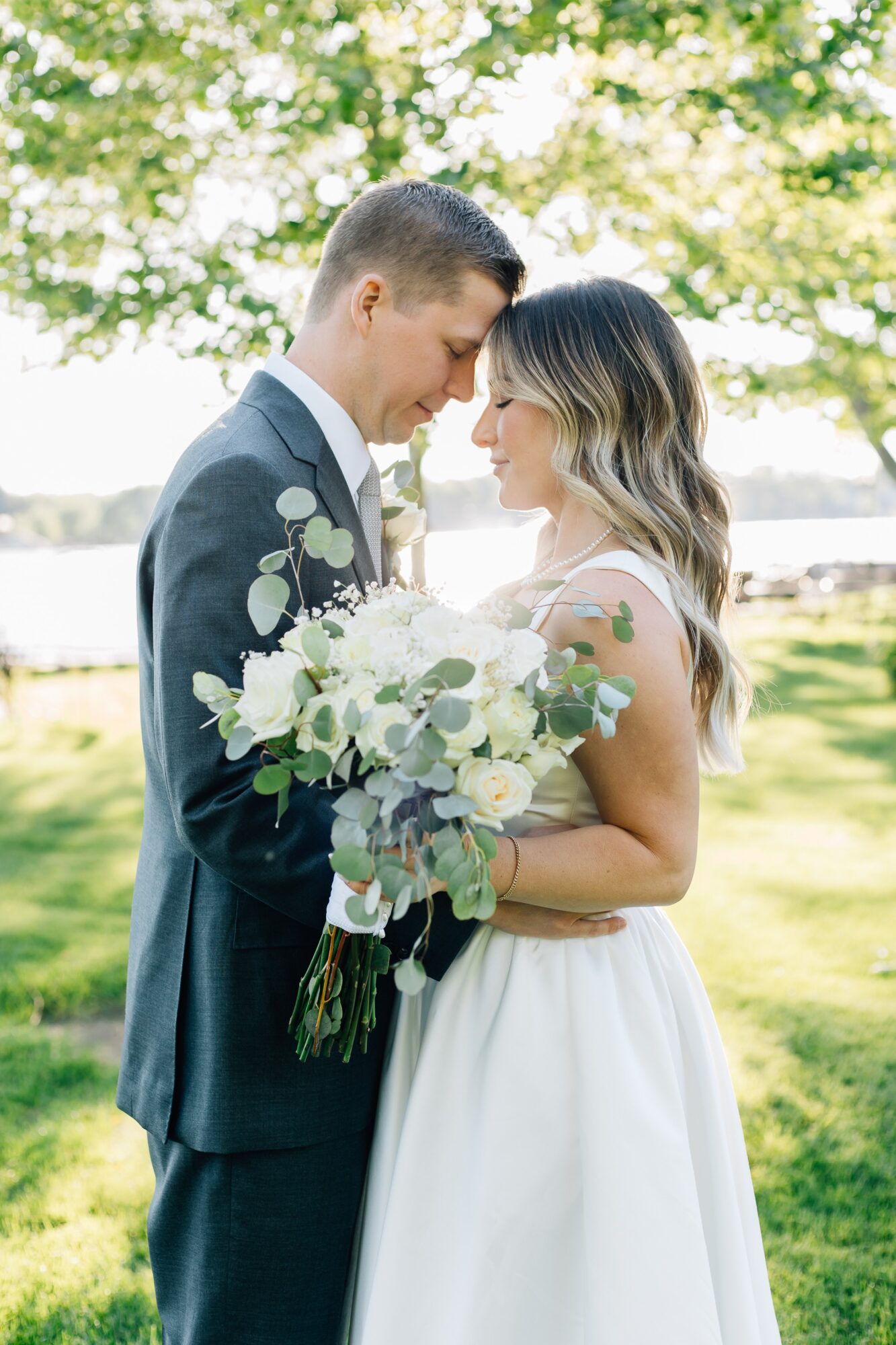 A couple in wedding attire stands close outdoors, holding a bouquet of white flowers, with trees and sunlight in the background.